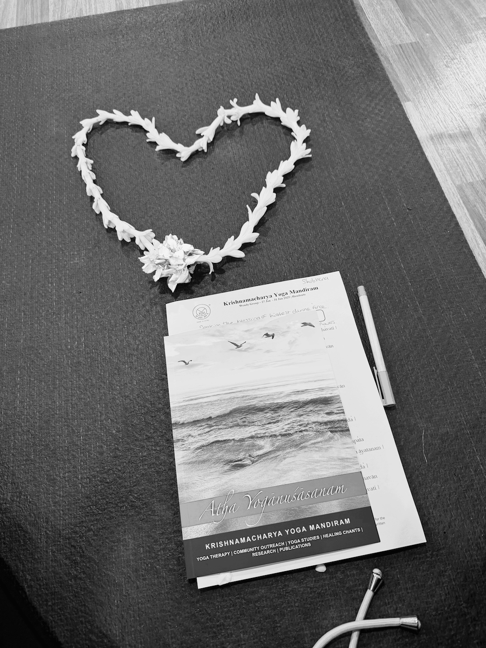 A black and white photo of a heart-shaped garland made of leaves on a dark textured surface. Below the garland, there are brochures for Krishnamacharya Yoga Mandiram, showing a beach scene with birds flying over the ocean, and some papers and a pen.