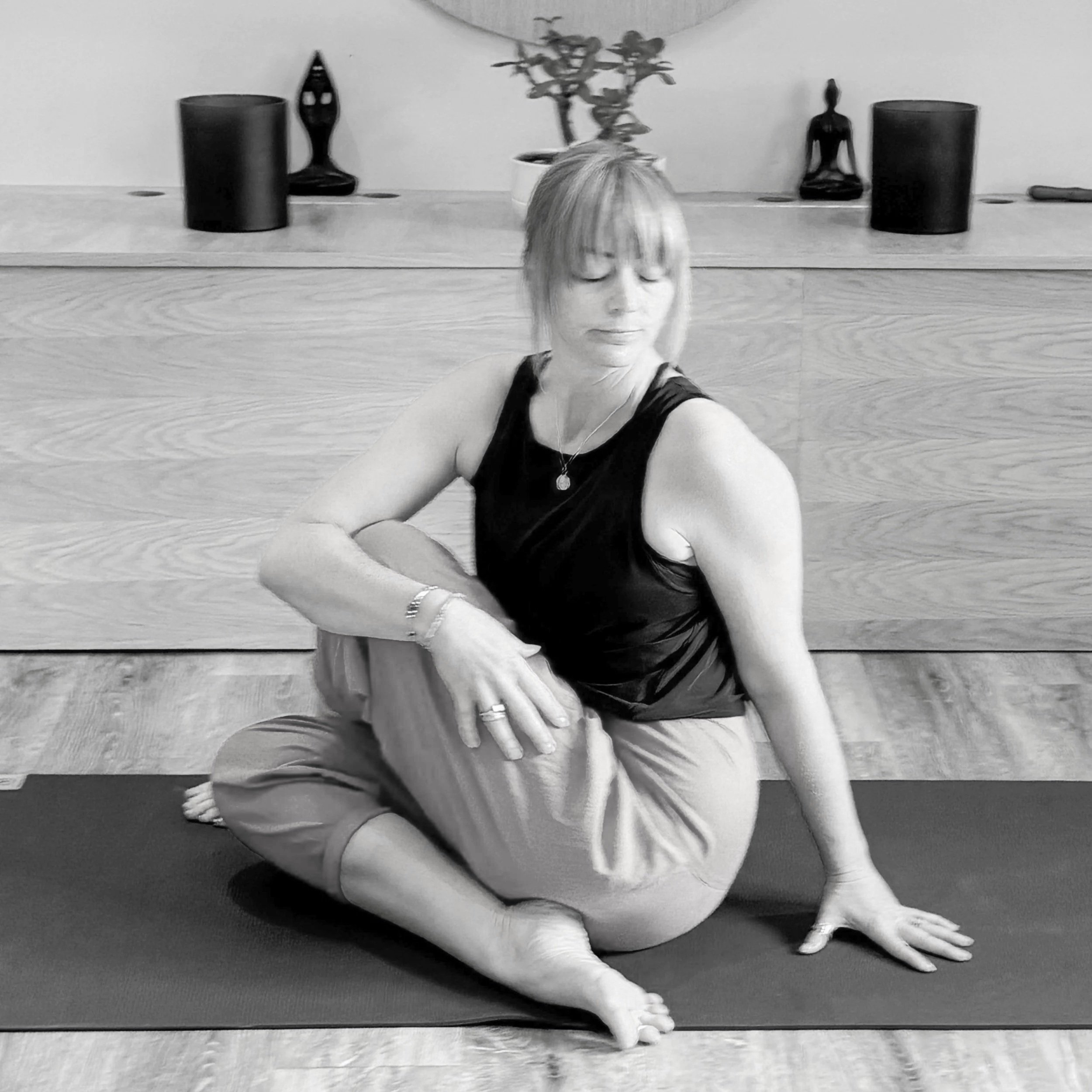 A woman with short hair and glasses practicing yoga on a mat indoors. She is sitting in a relaxed pose with her eyes closed, wearing a sleeveless top and jewelry, with decorative displays on a shelf behind her.