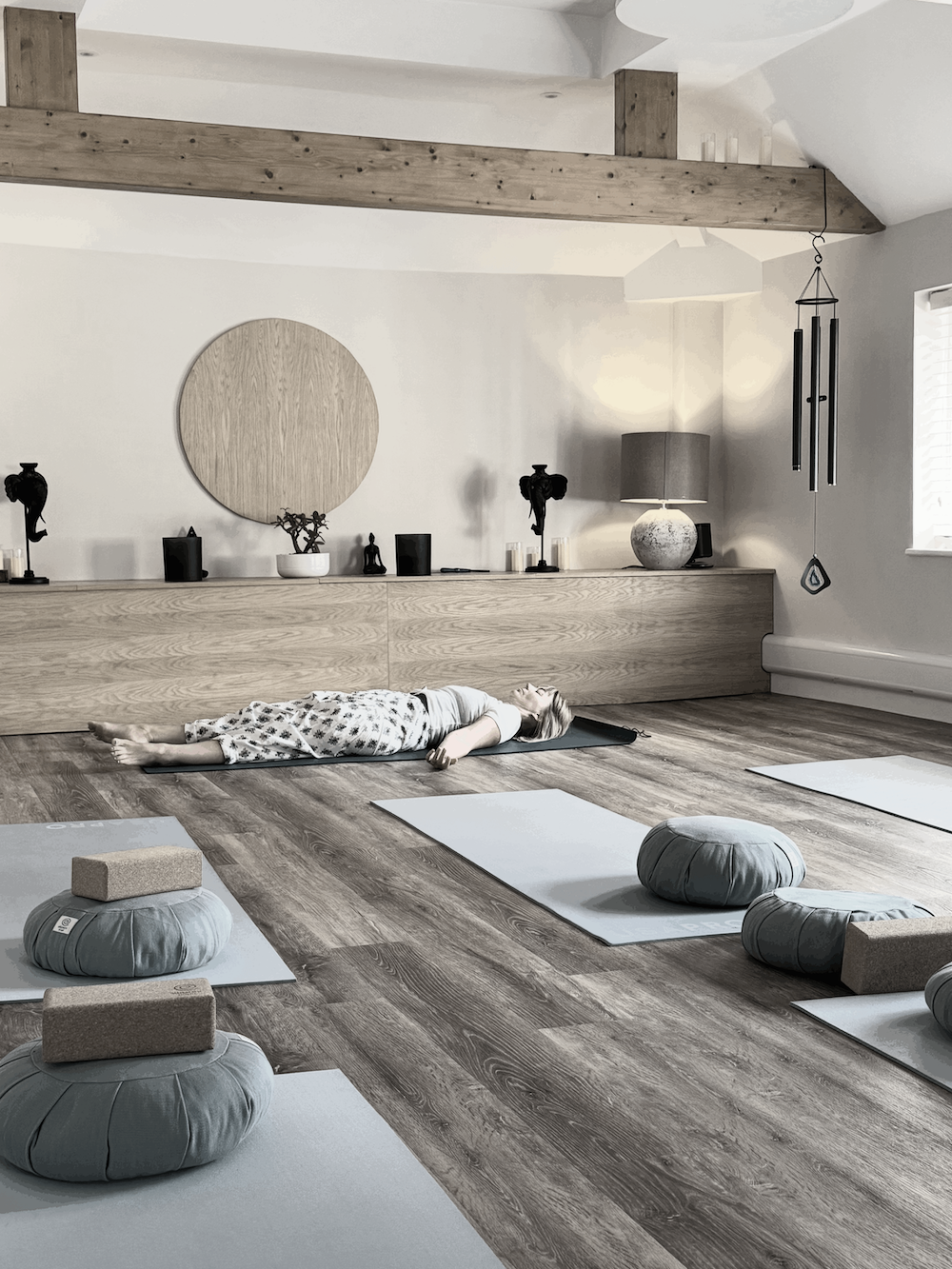 A person lying on a yoga mat in a peaceful, minimalistic room with wooden floors and neutral decor, surrounded by yoga cushions and blocks, with a sideboard and decorative items in the background.