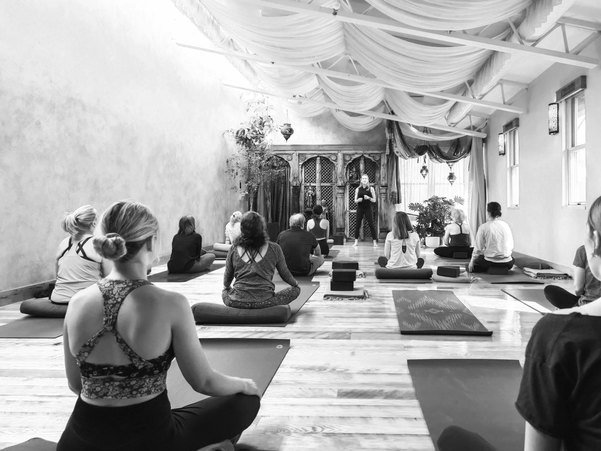 A group of women practicing yoga or meditation in a studio with draped fabric decor on the ceiling, large windows, and potted plants, seated on mats in a cross-legged position with their eyes closed or looking towards the instructor at the front.