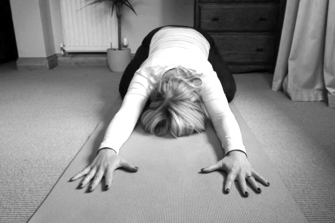 Woman practicing yoga in child's pose on a mat in a room with curtains, a potted plant, and furniture.
