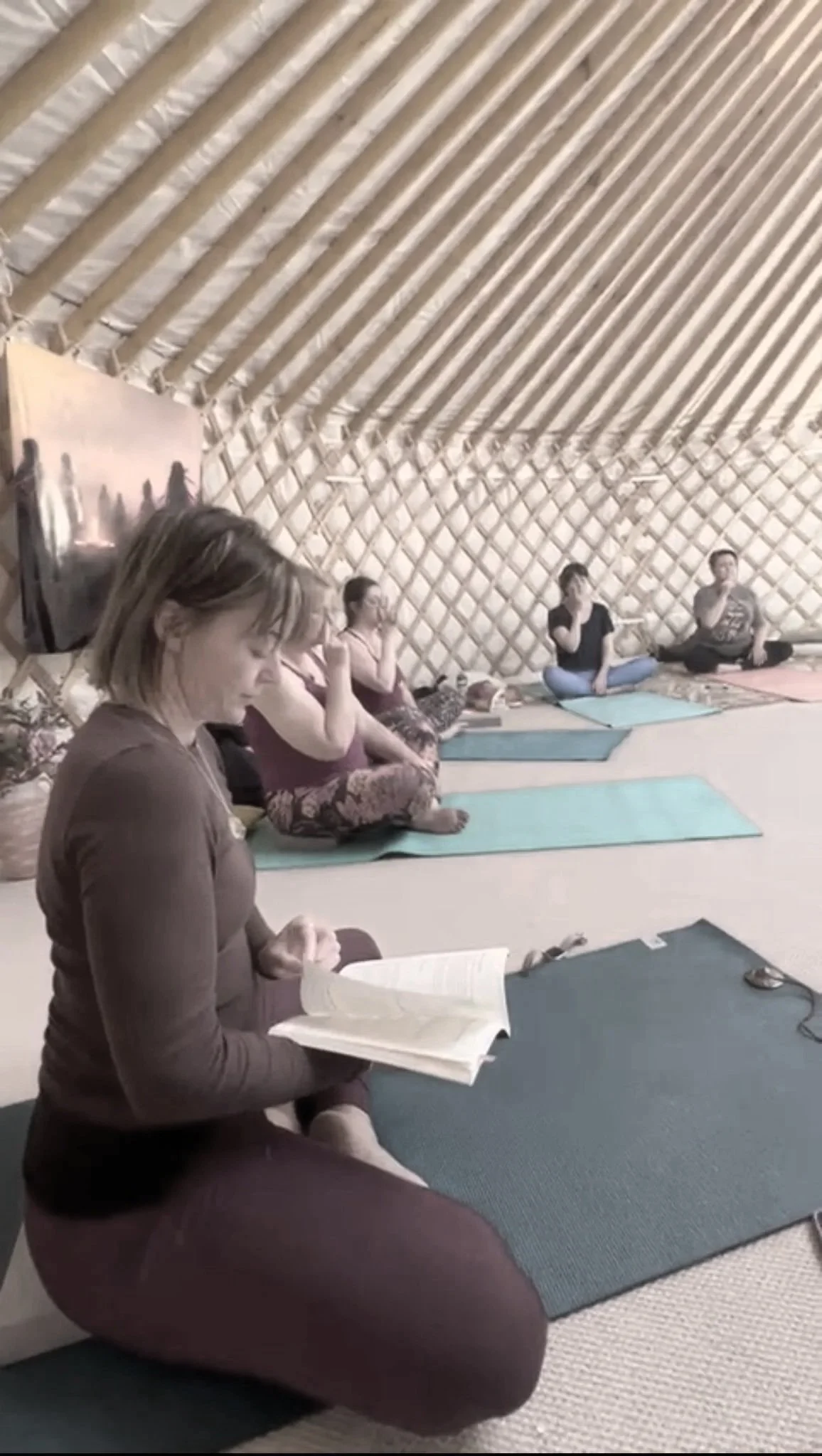 Women participating in a yoga or meditation class inside a yurt, sitting on yoga mats with a book, with some participants in a cross-legged seated position.