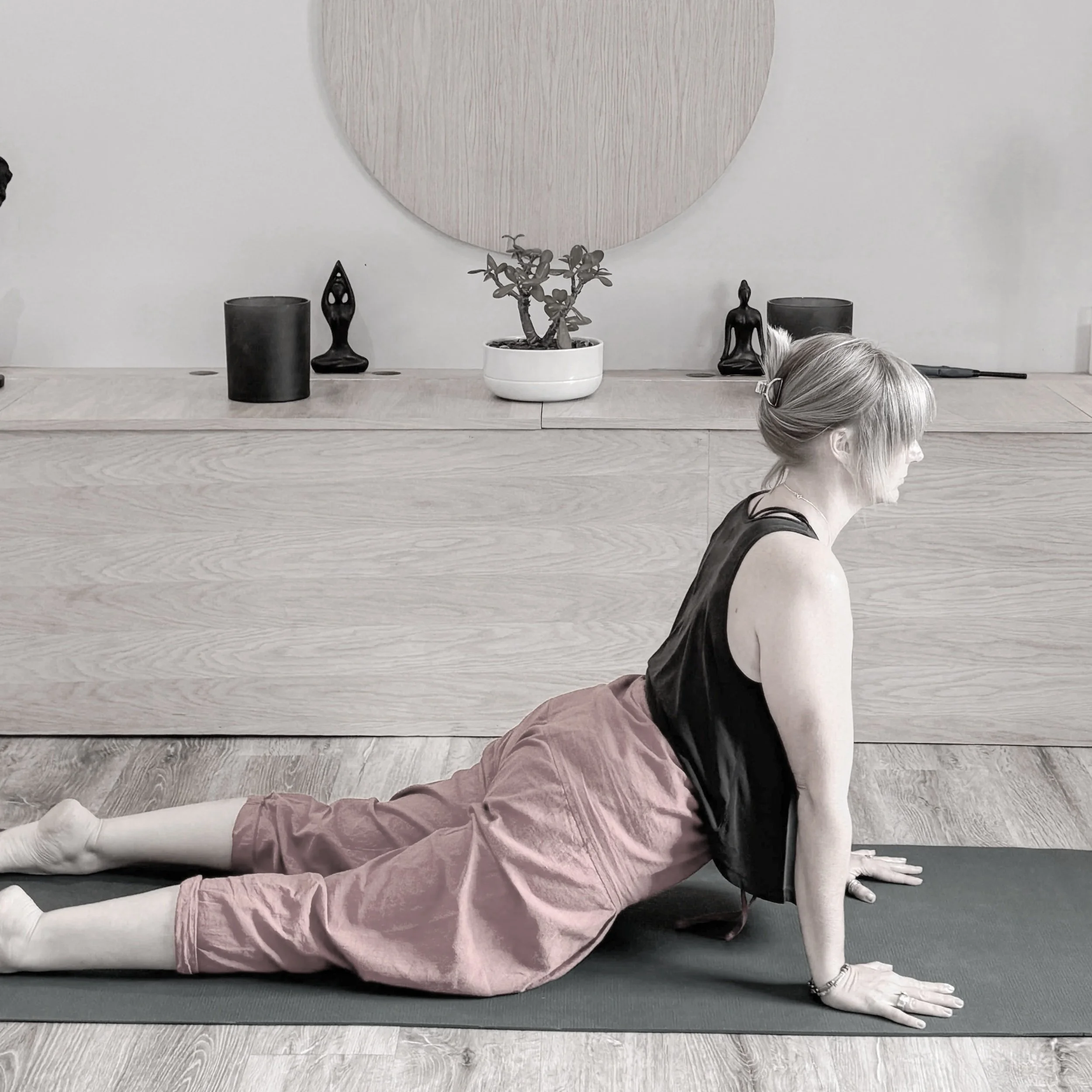 A person with silver hair practicing yoga on a black mat, in a side plank pose, wearing black top, pink pants, and white socks, with a white potted plant, black decorative items, and a large circular wall art piece behind them.