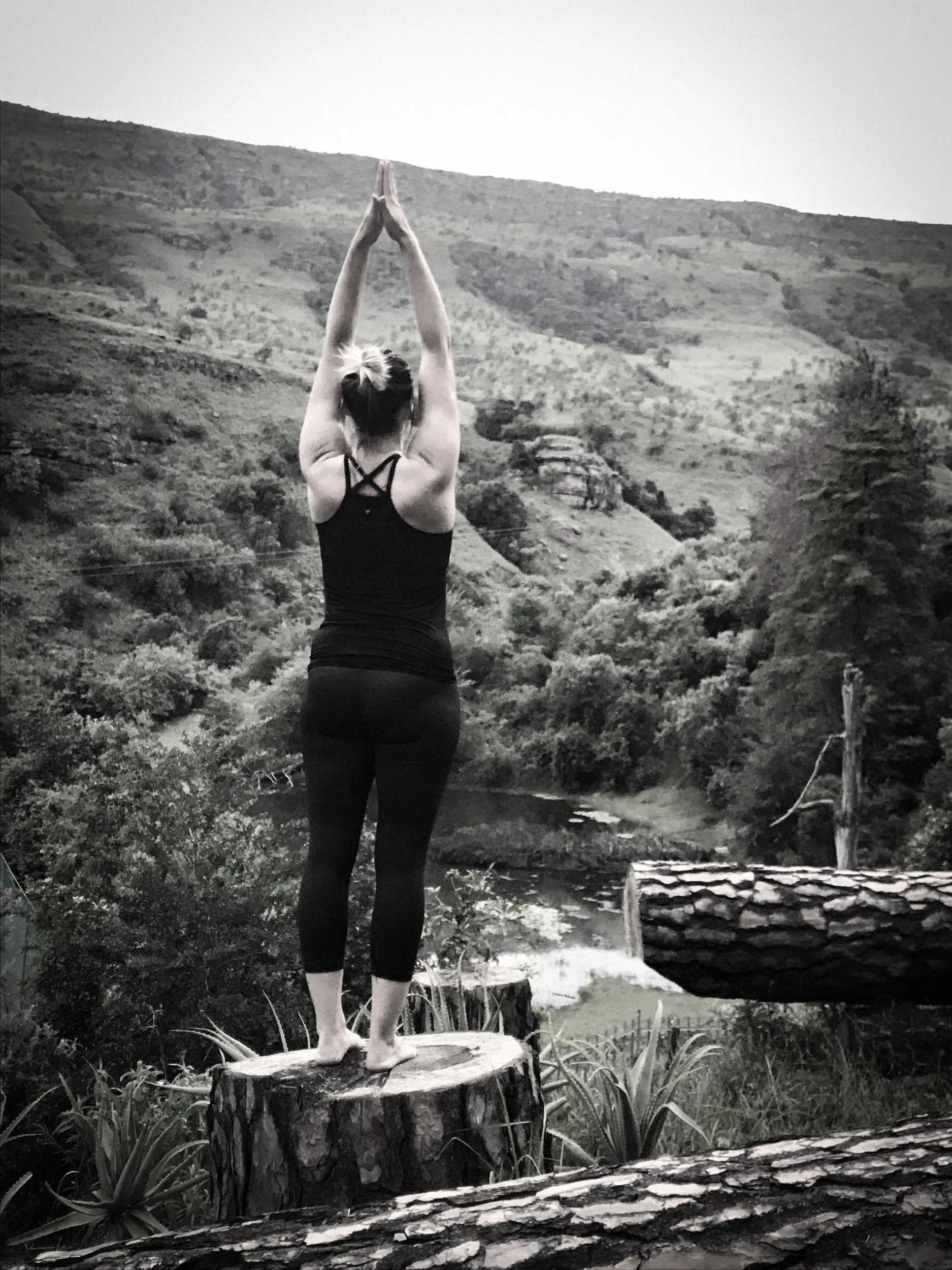 A woman practicing yoga outdoors on a tree stump in a mountainous landscape with trees and water, seen from behind in black and white.