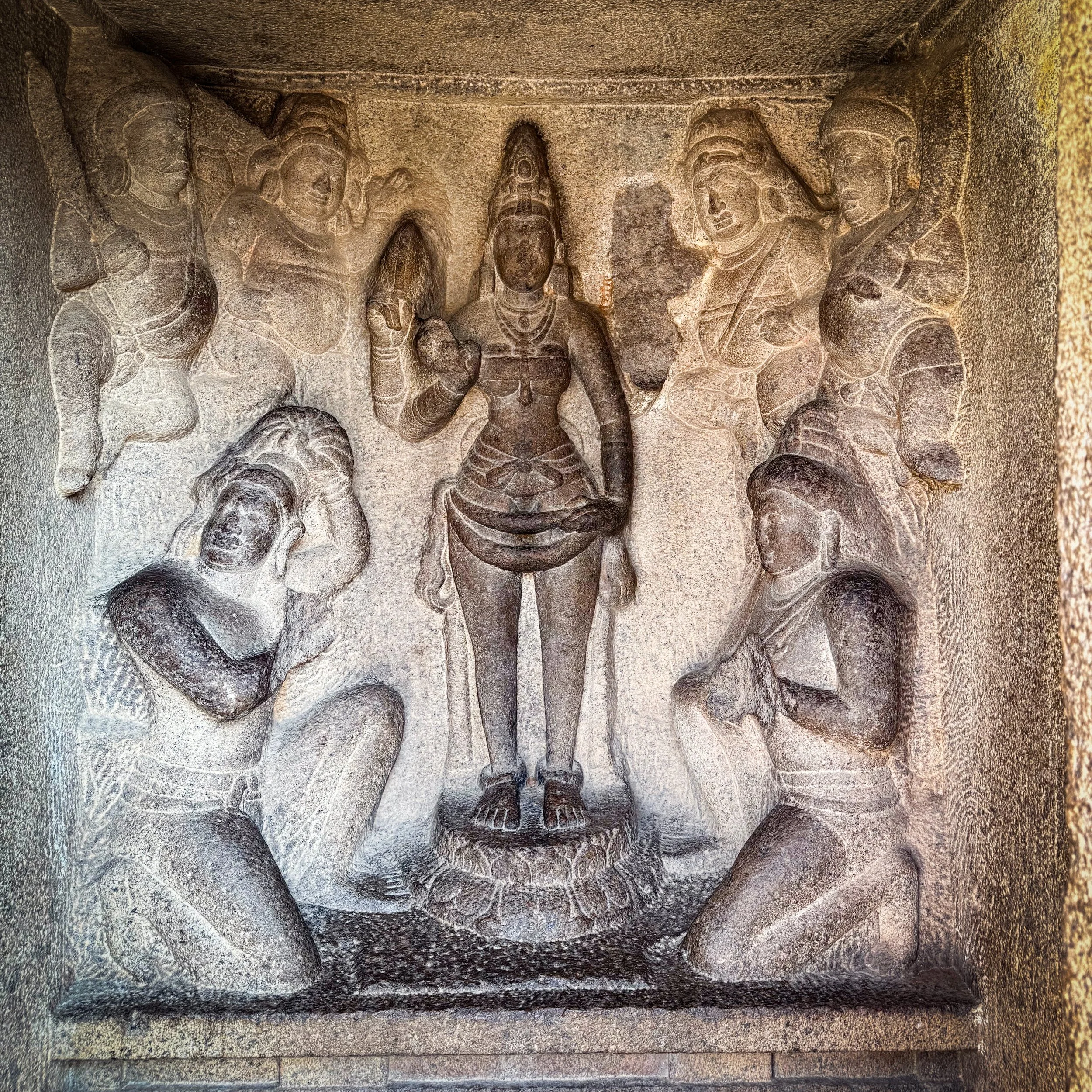 Ancient stone carving depicting a central figure adorned with a tall crown or headdress, surrounded by four other figures with headgear, all in a seated or standing posture, carved into a rough stone surface.
