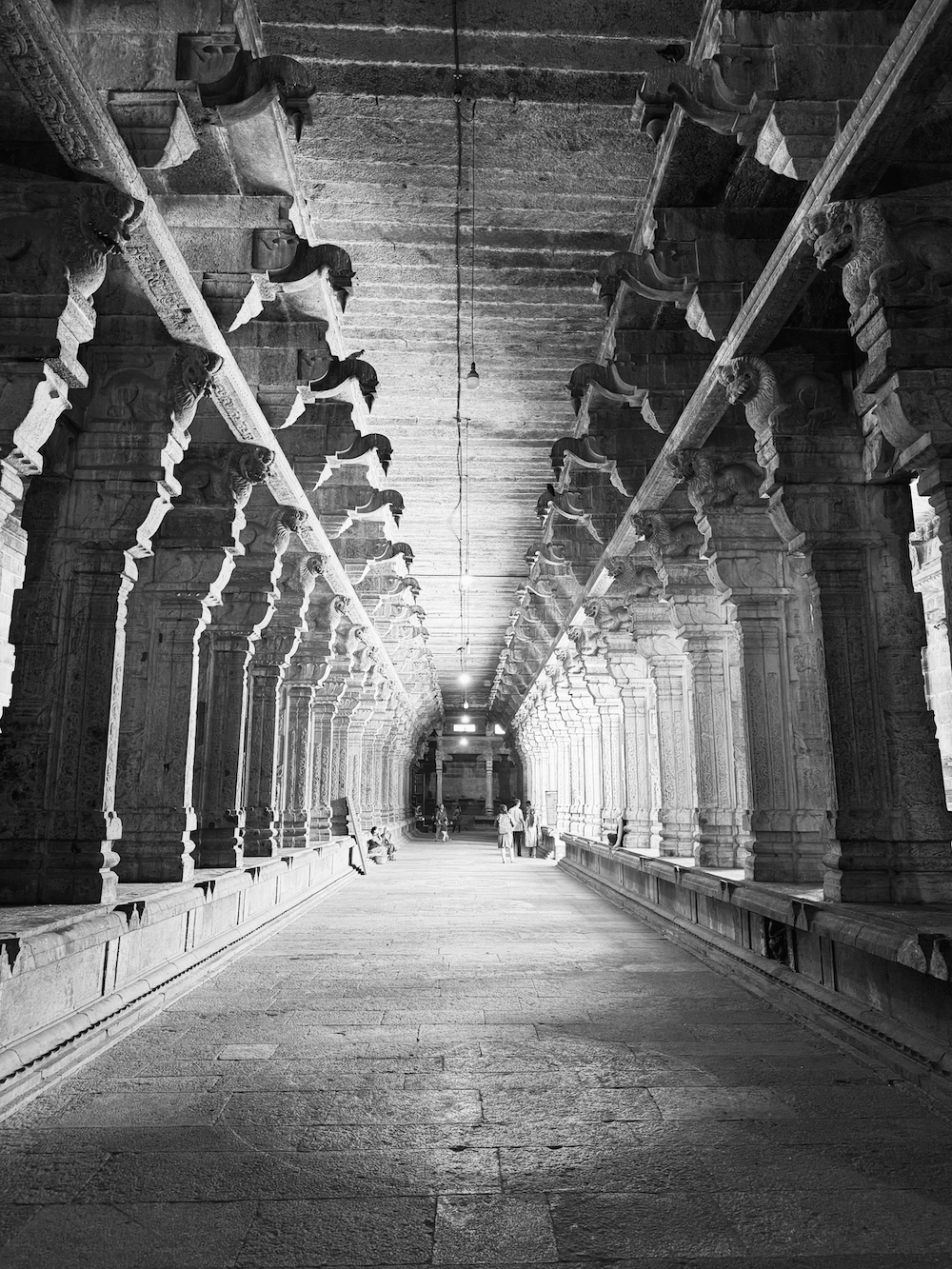 A black and white photo of an ancient temple corridor with intricately carved pillars and statues, with a few people walking and sitting along the sides.
