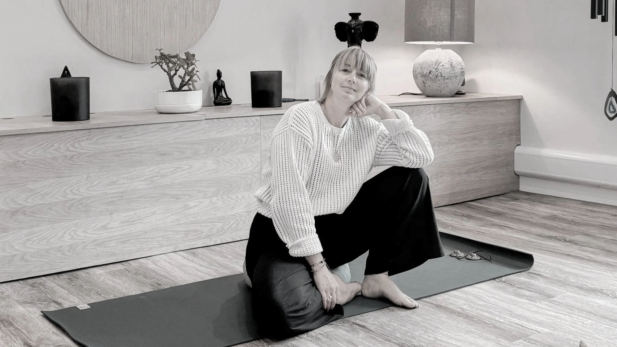 A woman doing yoga on a black mat indoors, sitting in a cross-legged position with her head resting on her hand, dressed in a white sweater and black pants, with decorative items and a lamp on a wooden sideboard behind her.