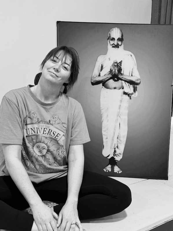 A woman sits cross-legged in front of a black-and-white image of an older man with hair tied back, holding a cloth and making a gesture with hands pressed together in a prayer-like pose.