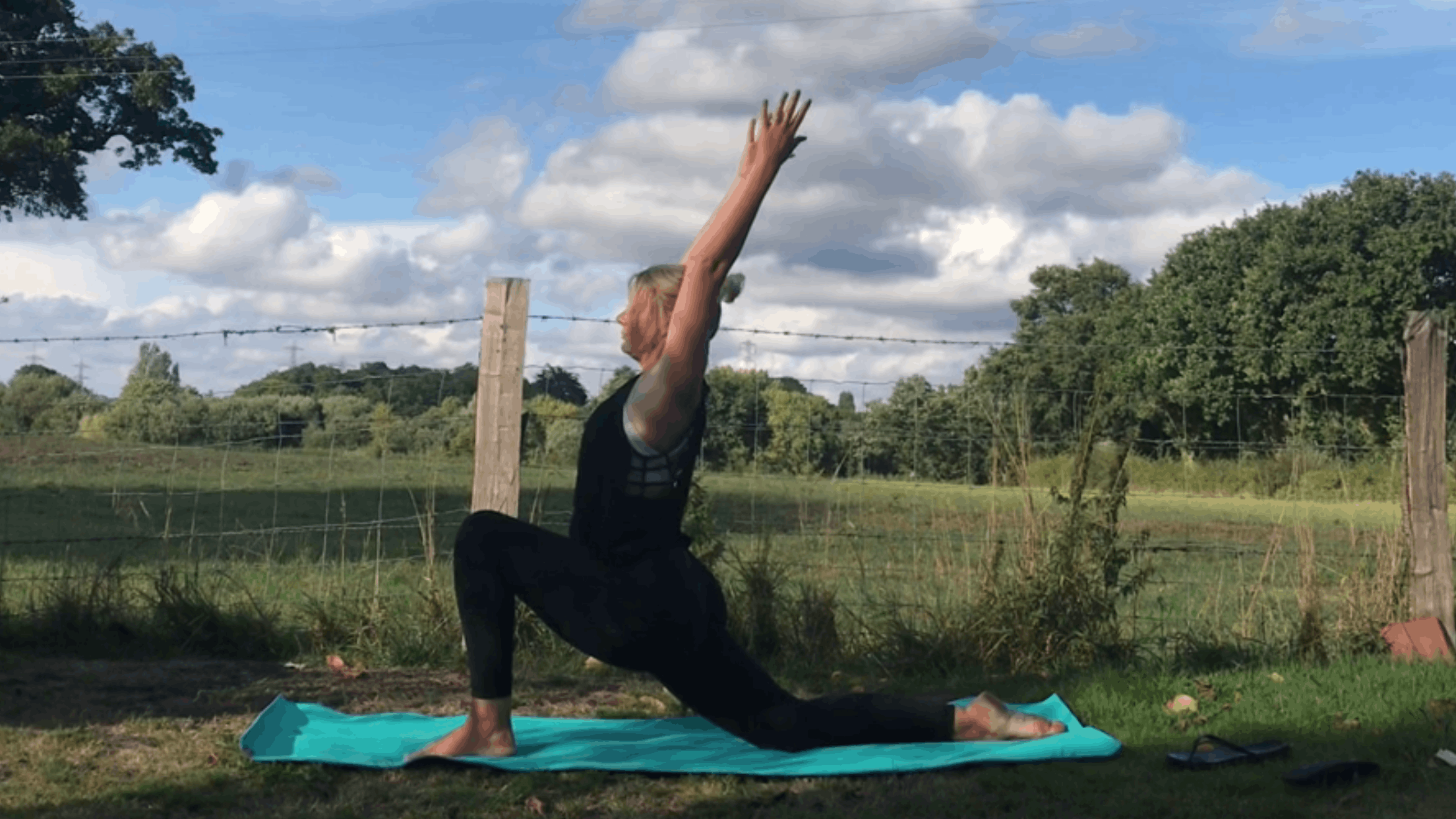 Person practicing yoga outdoors on a blue mat, performing a yoga pose with one hand on the mat and one arm raised towards the sky, in a grassy area with trees and a fence in the background, under a partly cloudy sky.