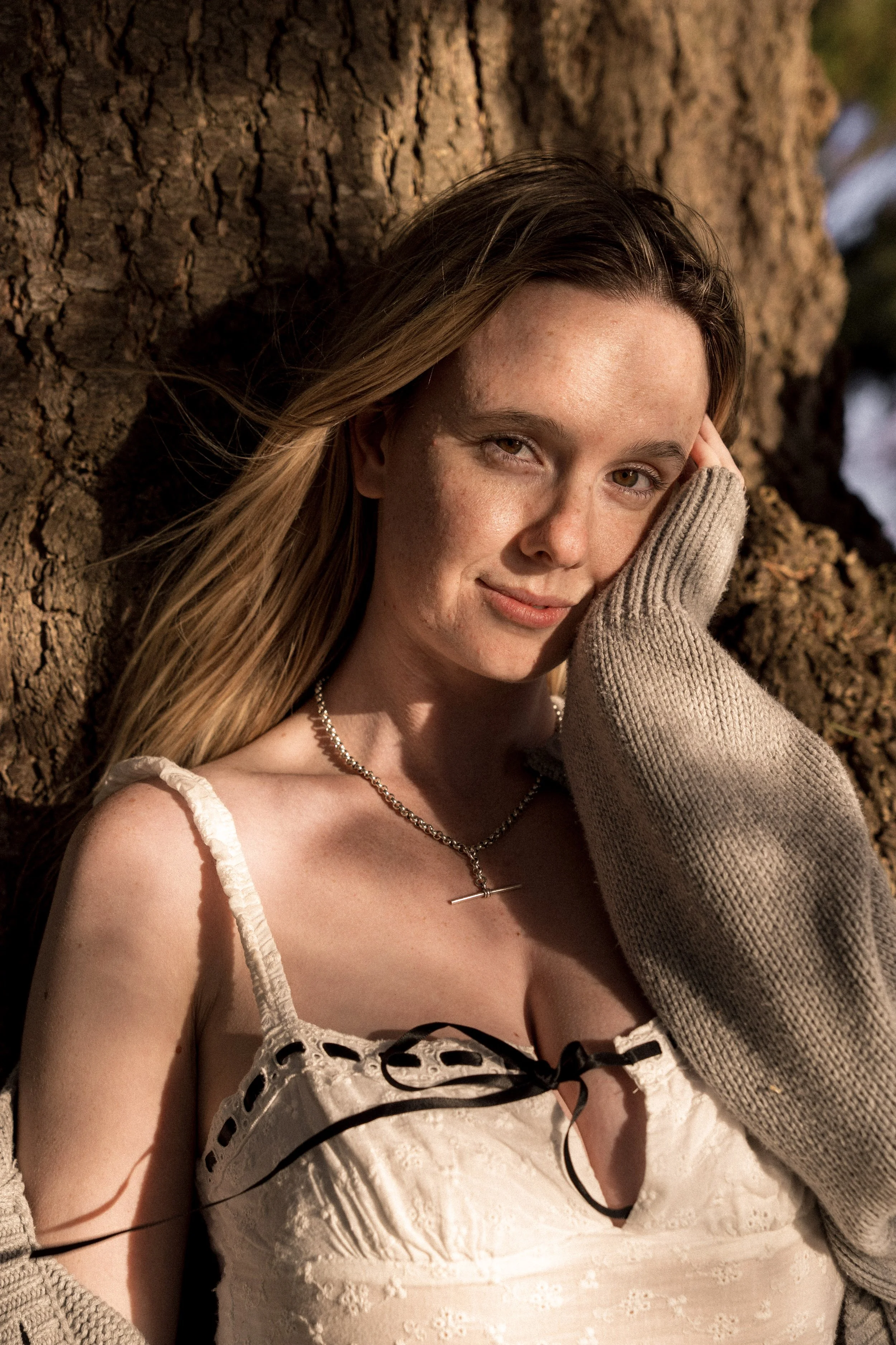 A young woman with long blonde hair and freckles leaning against a tree, smiling gently at the camera. She is wearing a white dress with black ribbon detailing, a silver necklace, and a beige sweater over her arm.