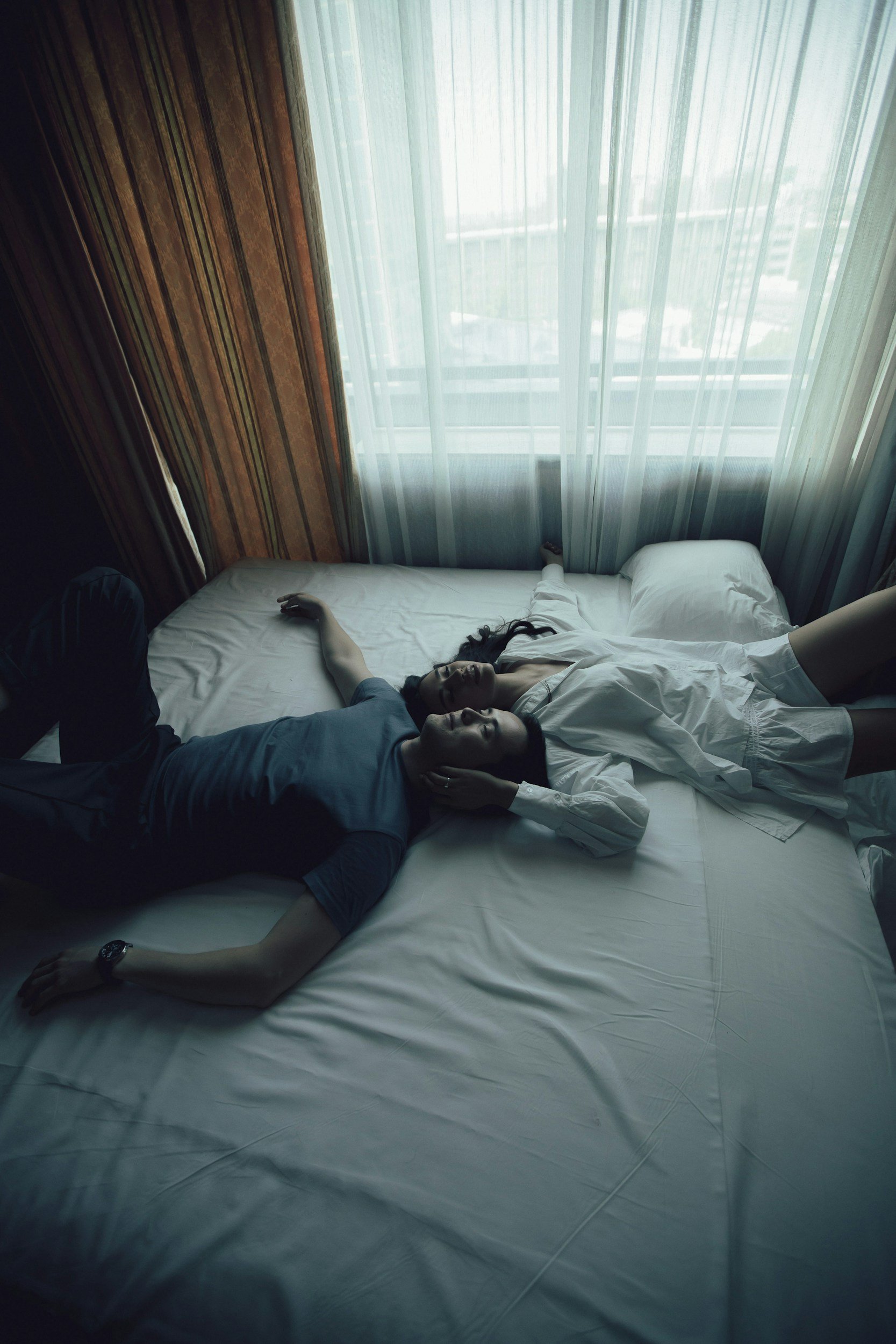 A high-angle, cinematic photograph of a man and a woman lying down together on a bed, looking upward in a relaxed, present state, representing physical connection and intimacy.