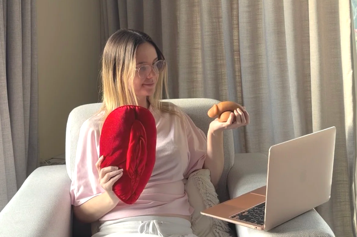 Somatic sex coach Michaela Tutty holding plush anatomical models during an online coaching session on laptop.