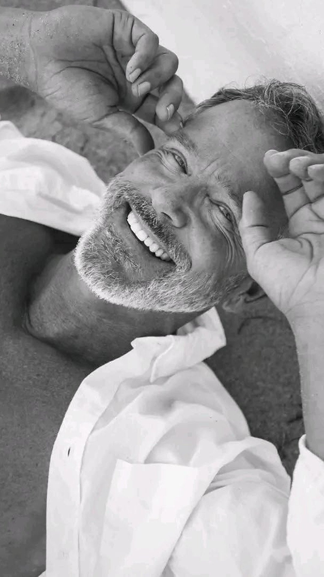 A smiling mature man lying down and resting his hand near his head, embodying relaxation, presence, and somatic sex coaching for men.