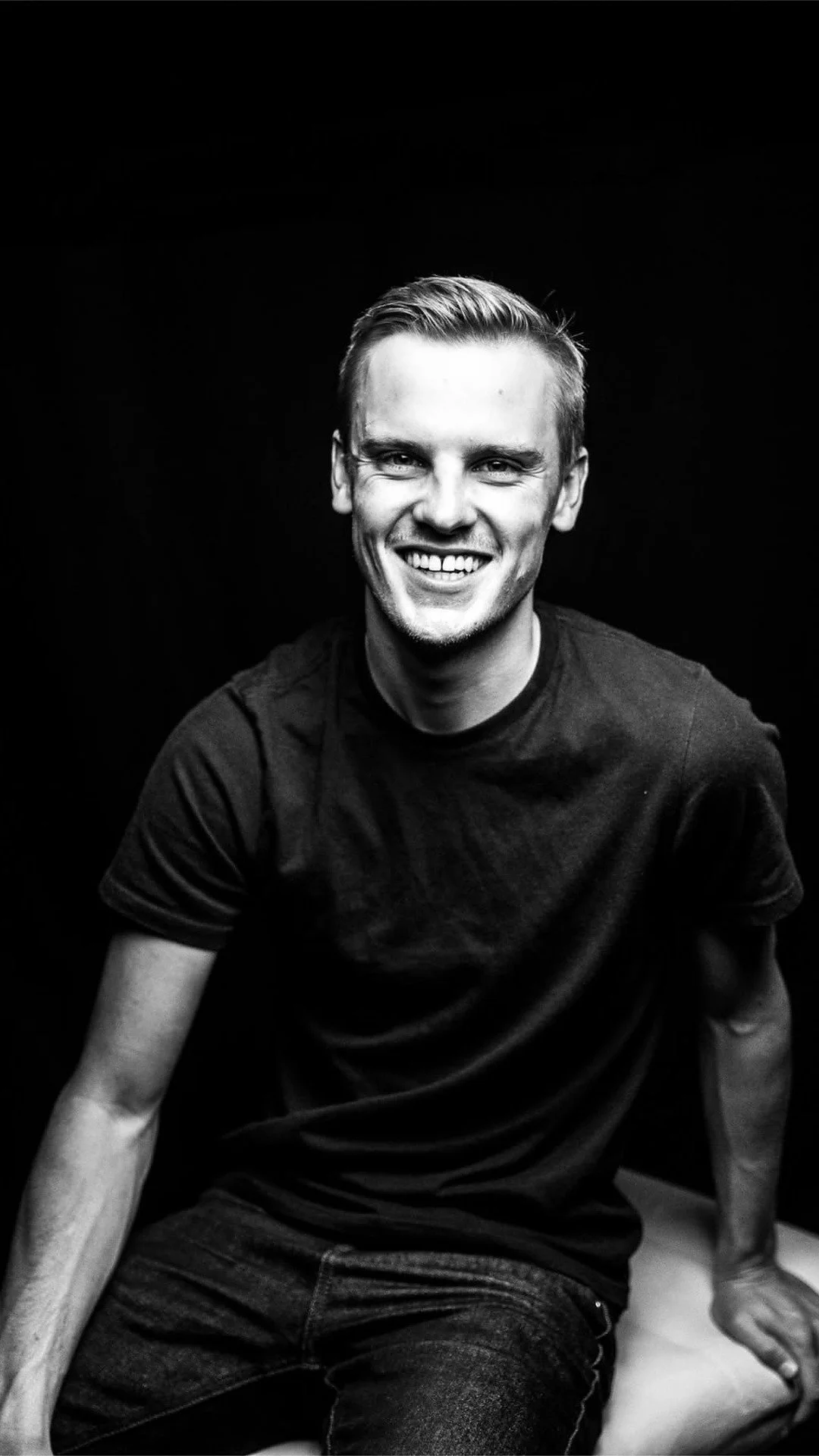 A man in a t-shirt sitting confidently and smiling at the camera, illustrating body awareness and relaxed masculinity.