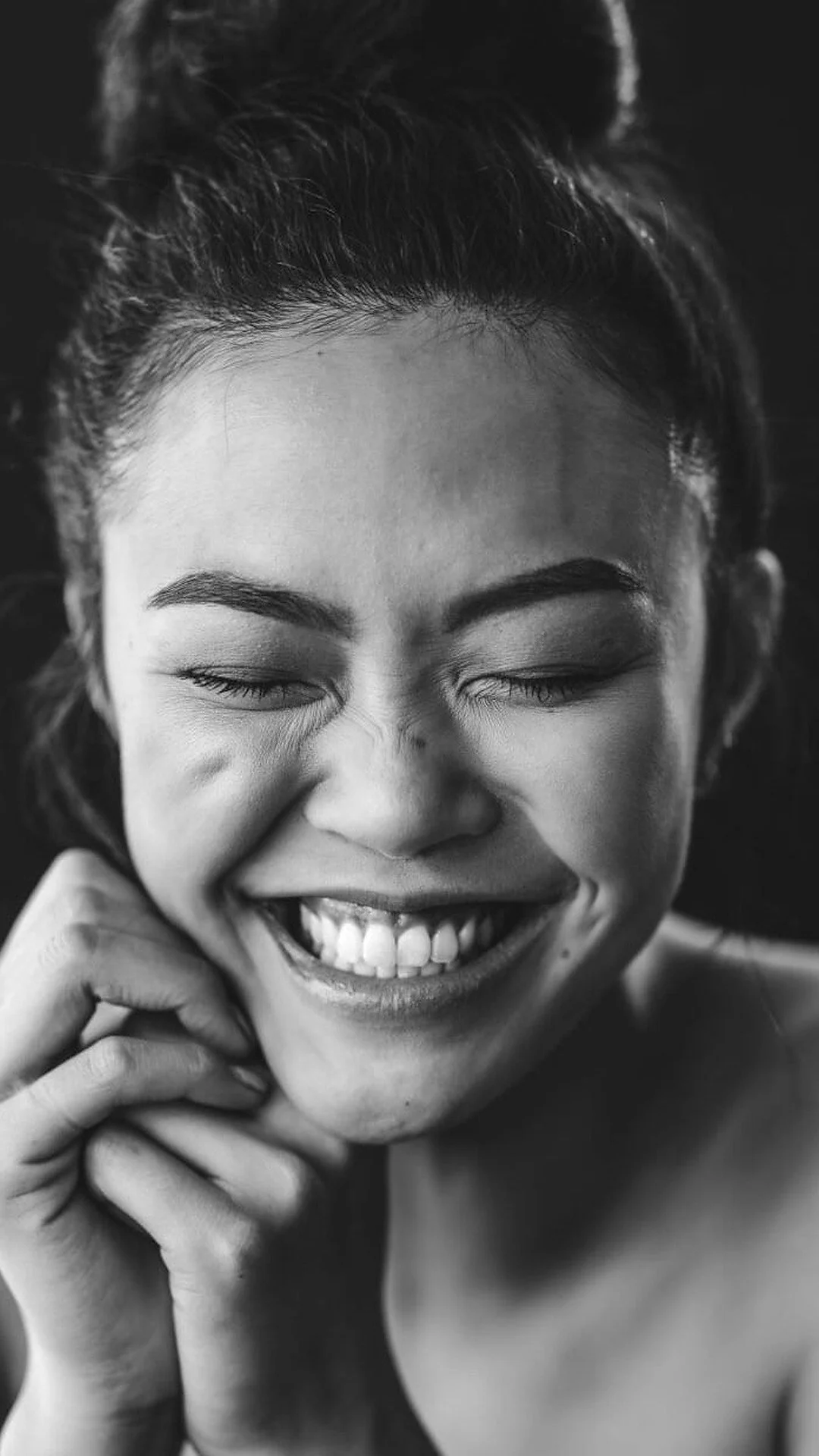 A smiling woman with her eyes closed holding her hands to her face in a candid moment of joy and body presence.