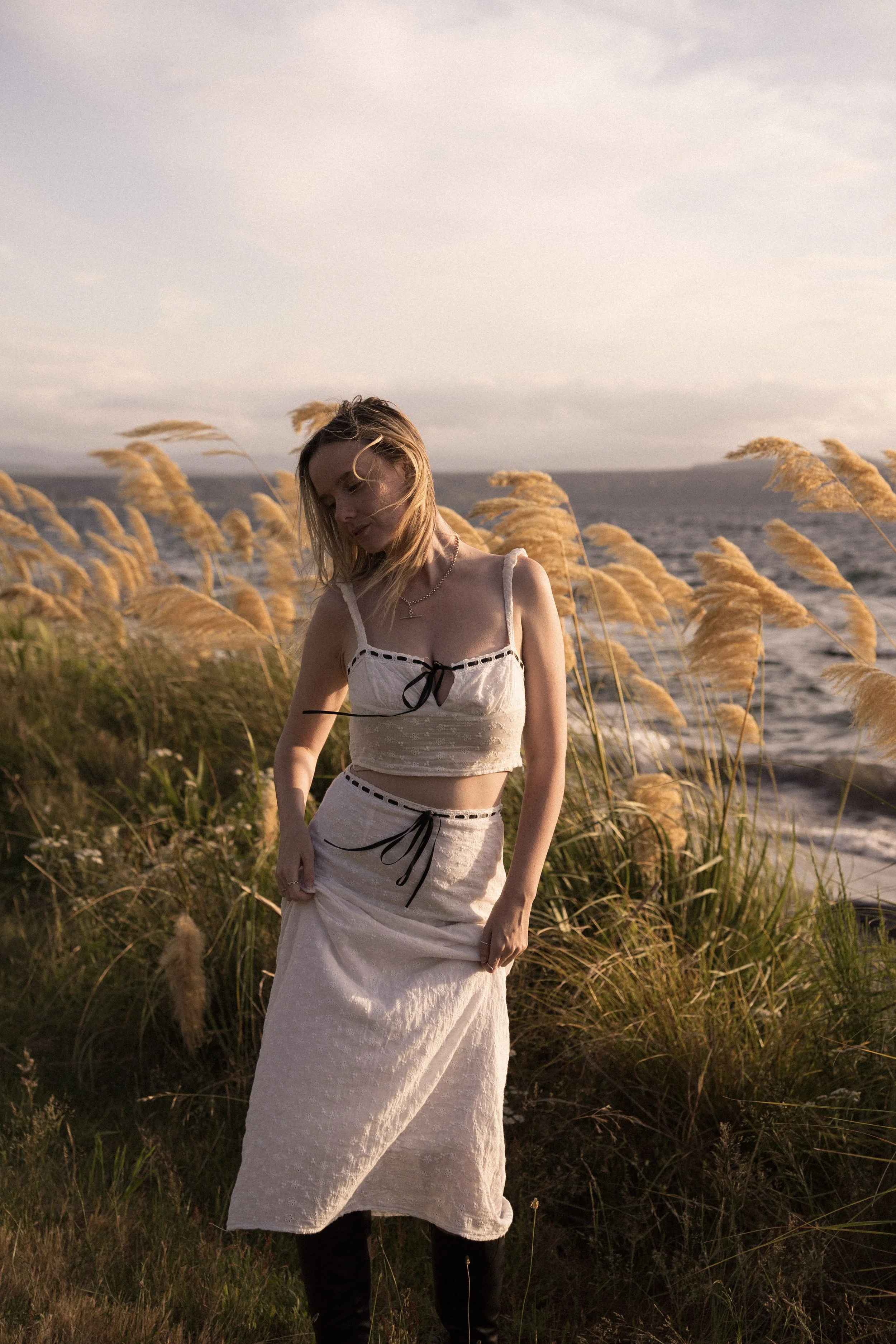 Michaela Tutty, a certified somatic sexologist in New Zealand, smiling in a white dress while standing in a grassy field alongside the scenic waters of Lake Taupō