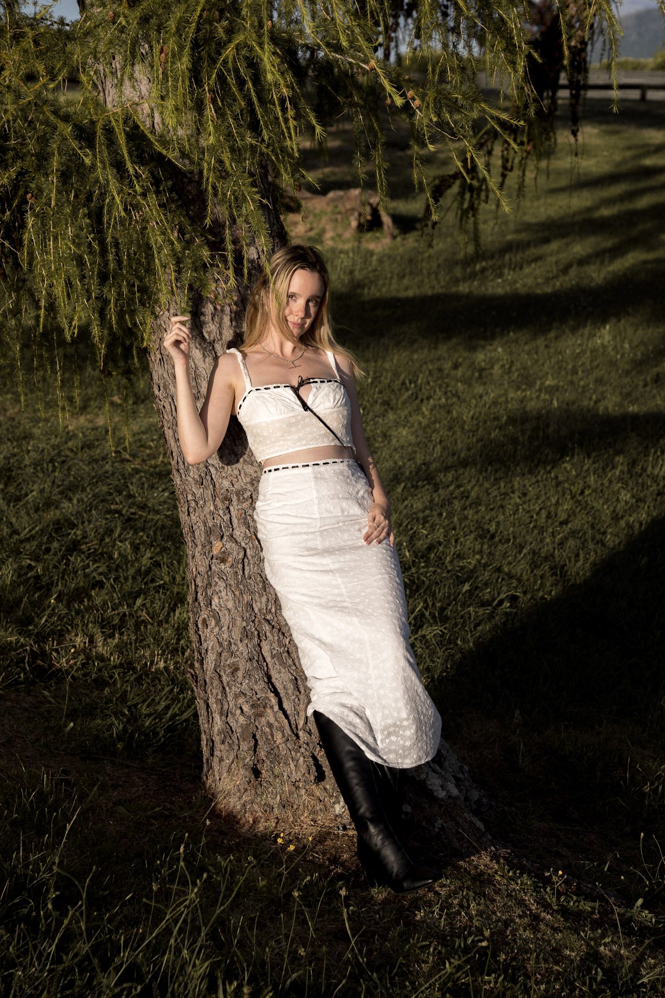 A woman in a white dress and black boots leaning against a tree, outdoors on a grassy area with sunlight and shadows.