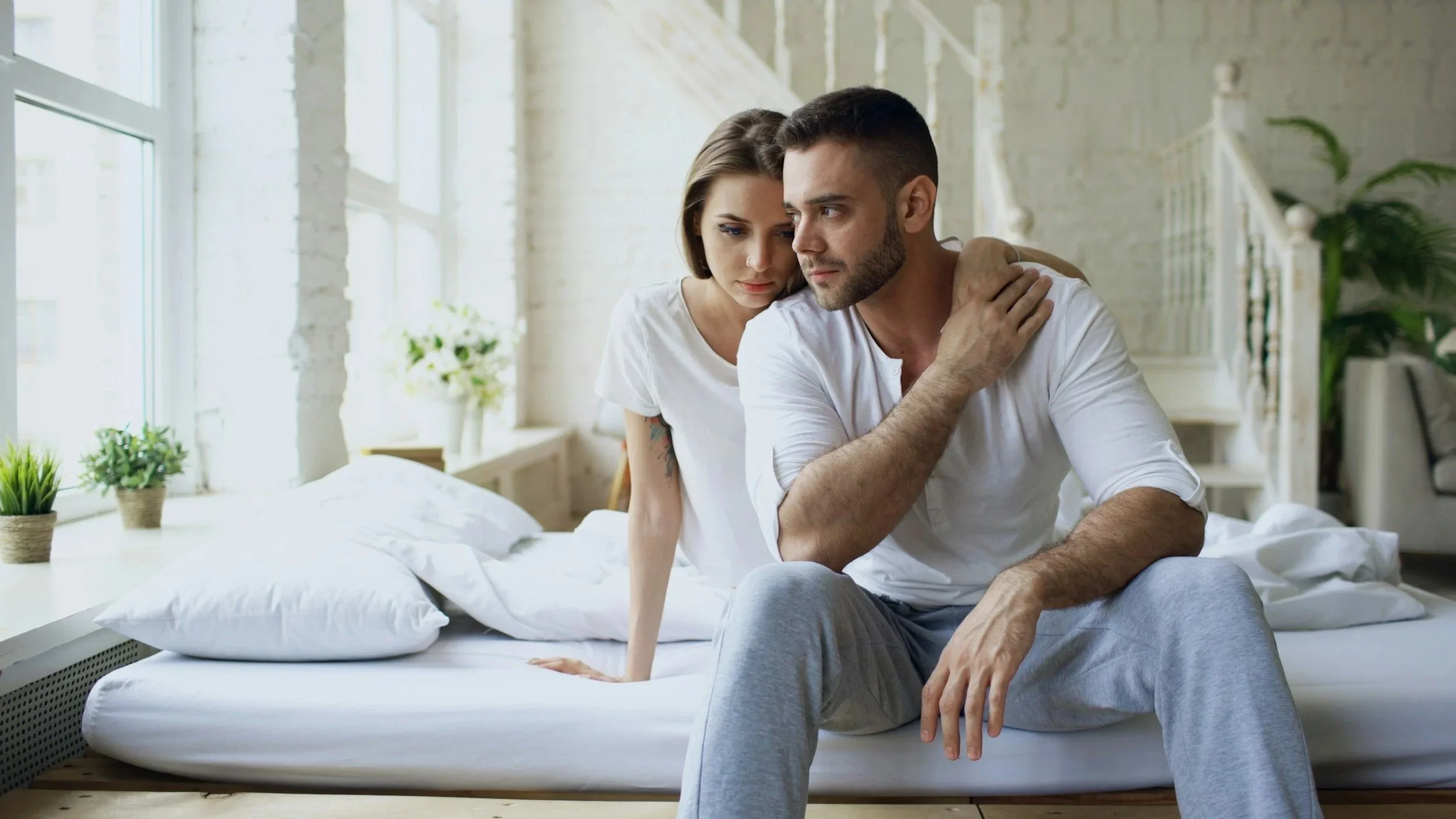 A thoughtful man and woman sitting on the edge of a bed looking in different directions, illustrating emotional closeness but physical disconnect in a long-term relationship.