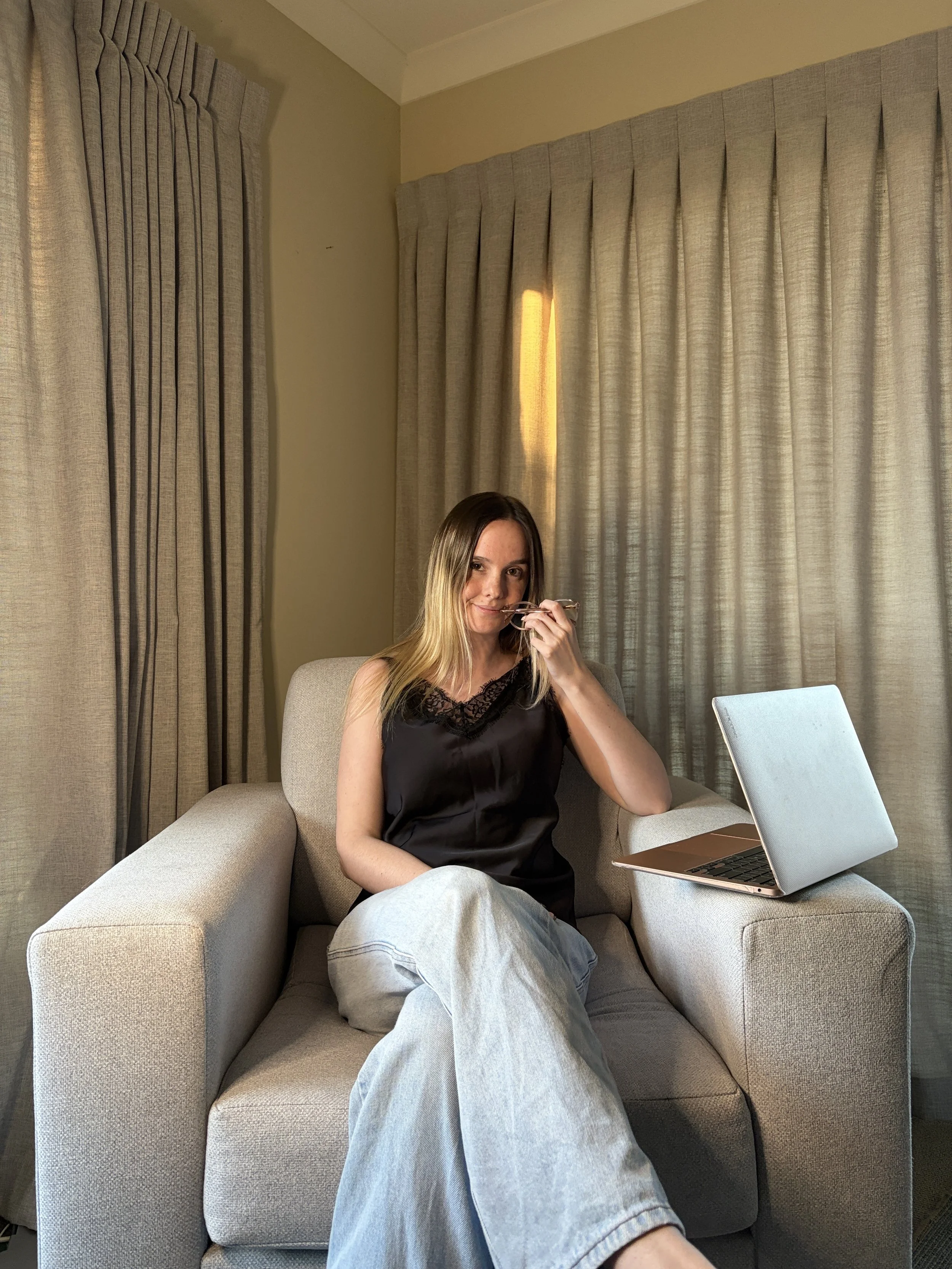 Michaela Tutty, somatic sexologist in Taupō, sitting in an armchair with a laptop in a cozy home office setting.