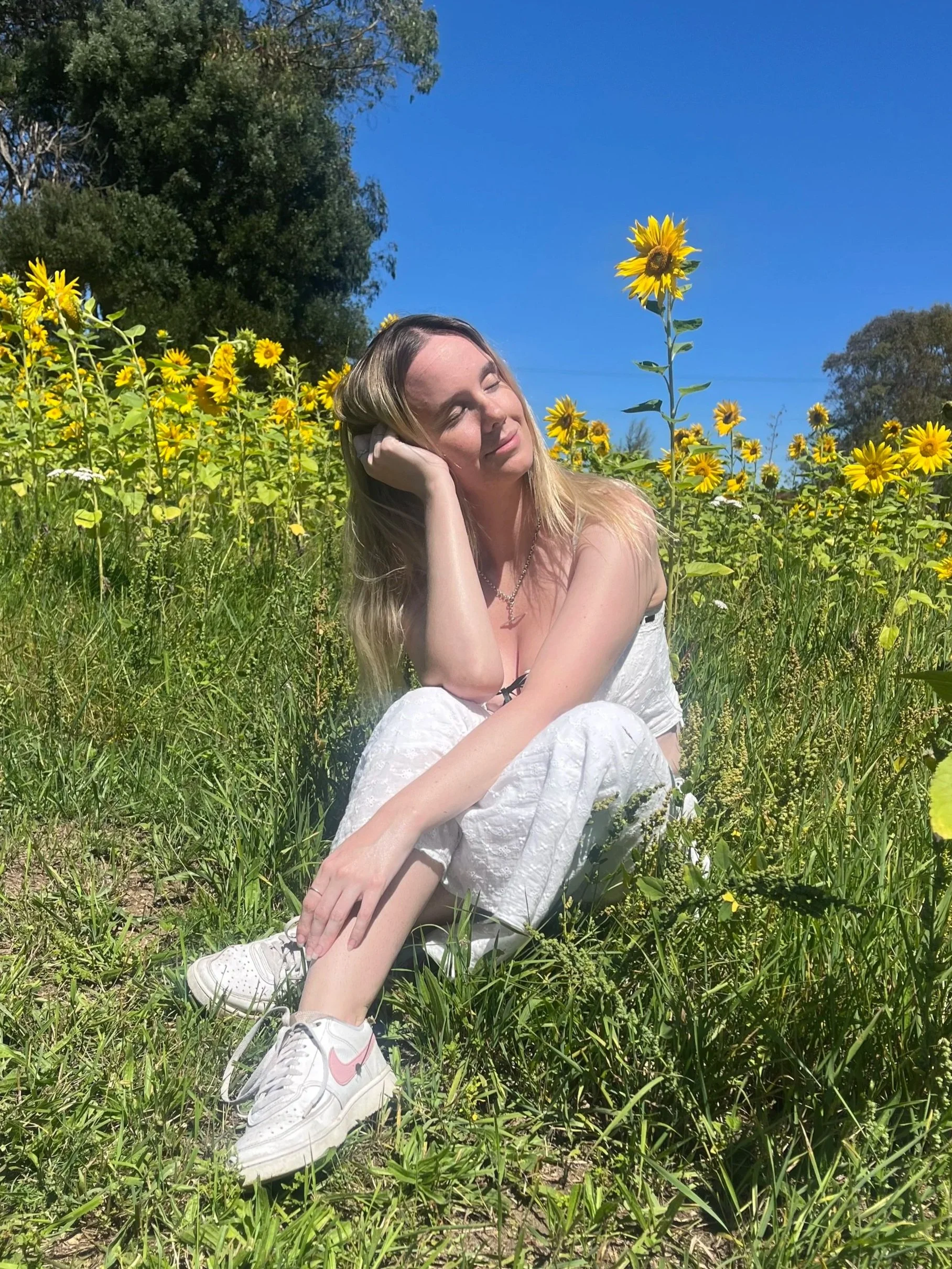Michaela Tutty, a somatic sex coach and sexologist, sitting in a sunflower field in New Zealand, reflecting a grounded and natural approach to intimacy and embodiment.