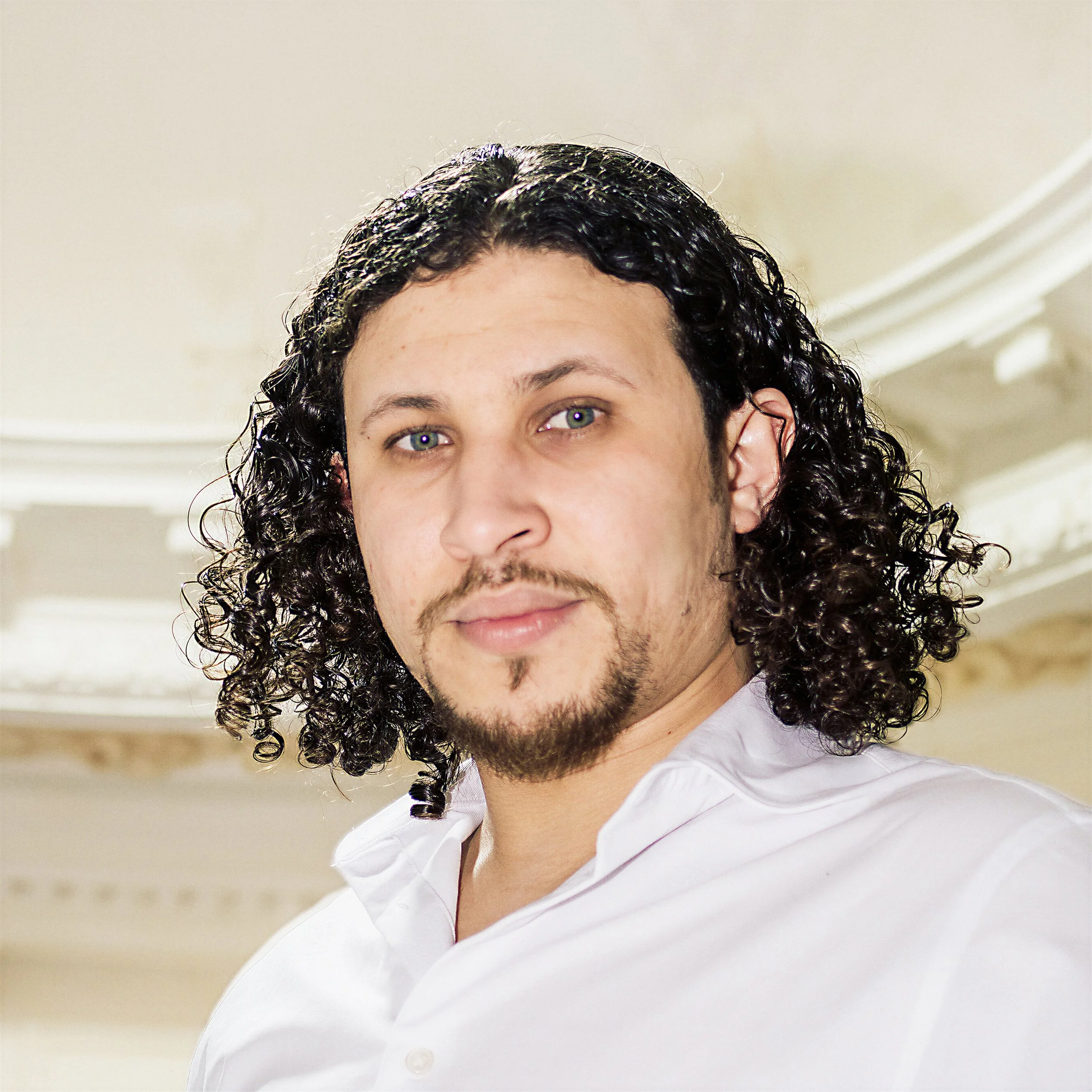 Portrait d'un homme avec cheveux bouclés, barbe et chemise blanche, dans une salle élégante.