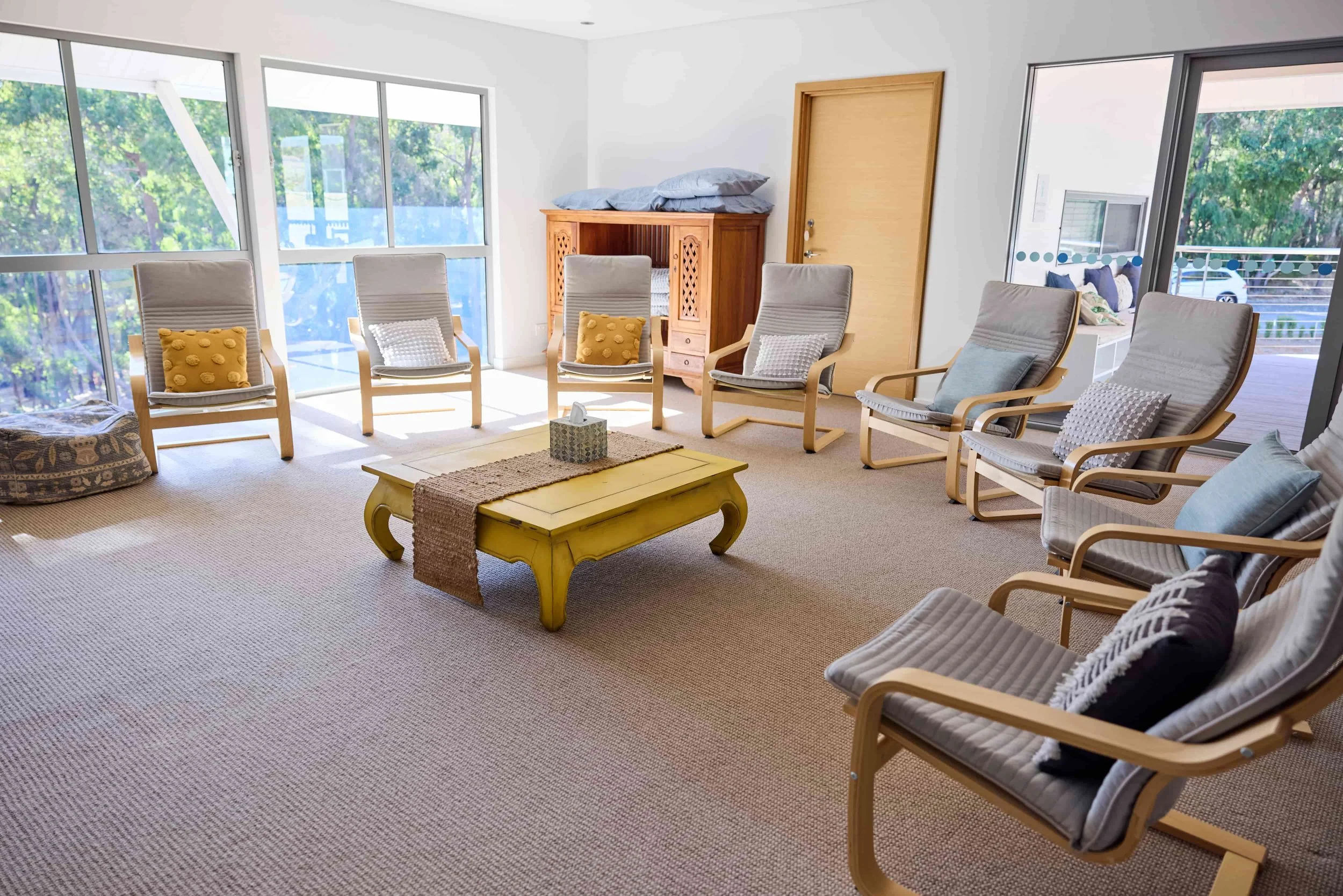 A bright living room with multiple armchairs arranged around a yellow coffee table, large windows and glass doors letting in natural light, with pillows on the chairs, and a wooden cabinet against the wall.