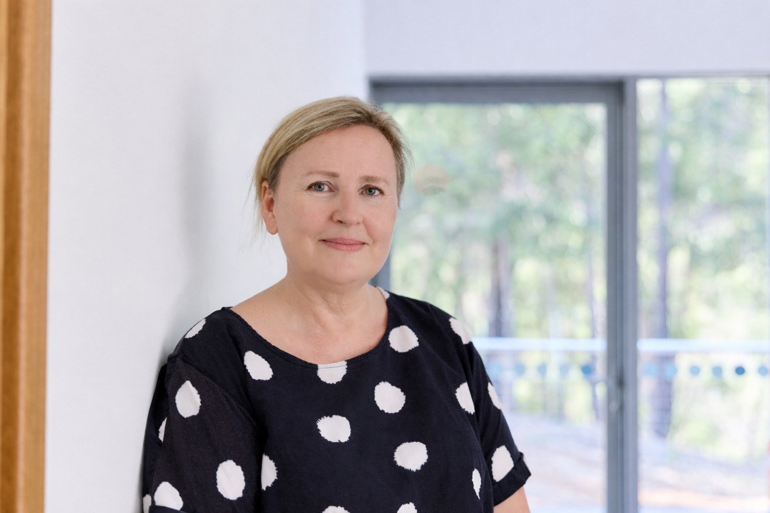 Owner of mindingu, Lorna, wearing a black top with large white polka dots, standing indoors near a white wall with a large window in the background showing trees.