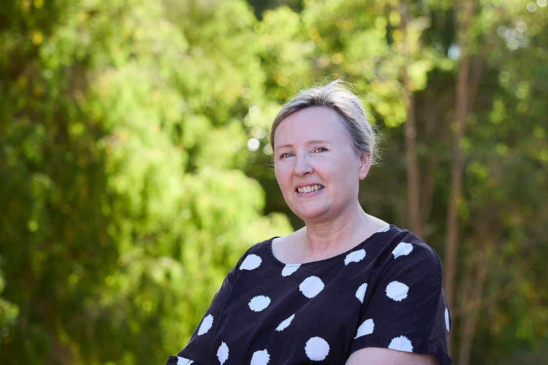 Owner and lead facilitator, Lorna, woman with short gray hair smiling outdoors, wearing a black shirt with white polka dots, in front of a background of green trees.
