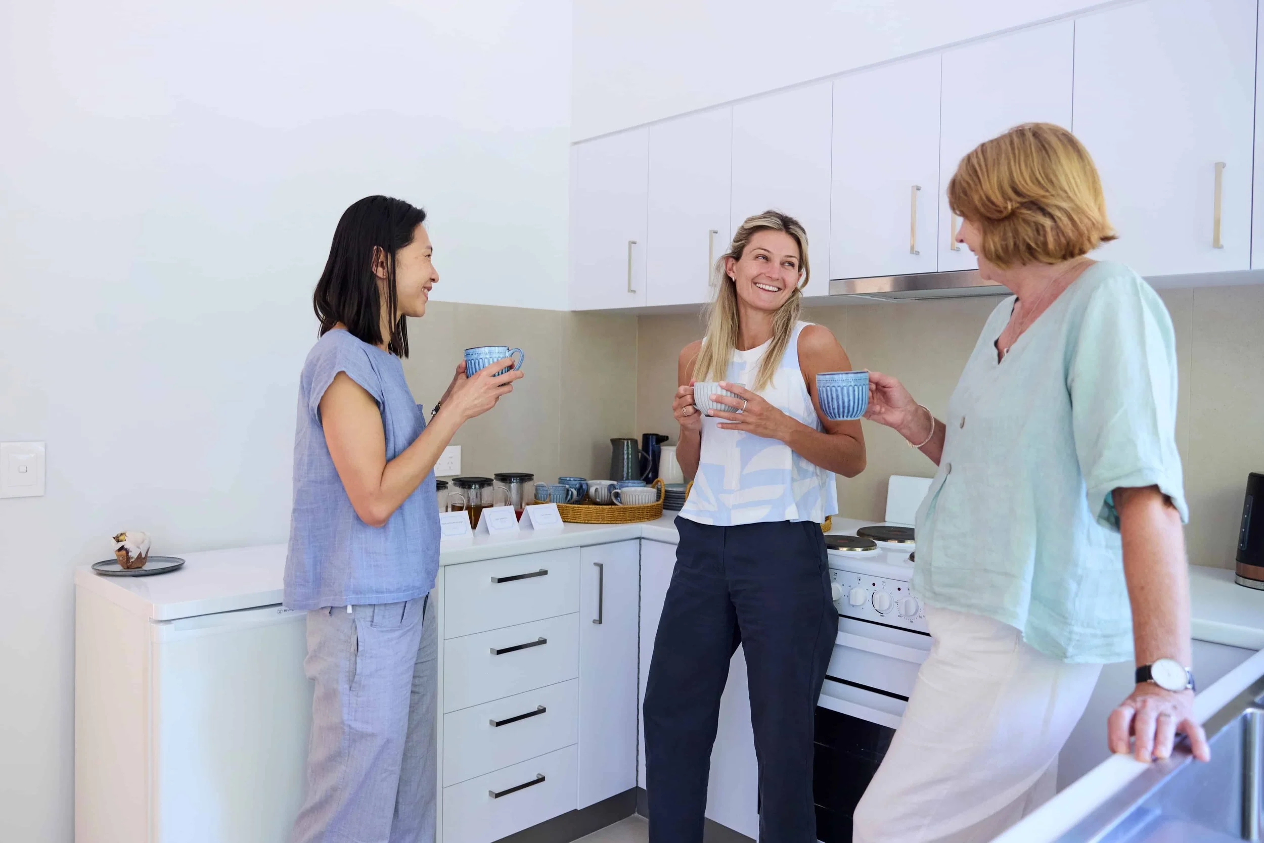 Three women in a kitchen chat and smile while holding cups of coffee or tea.