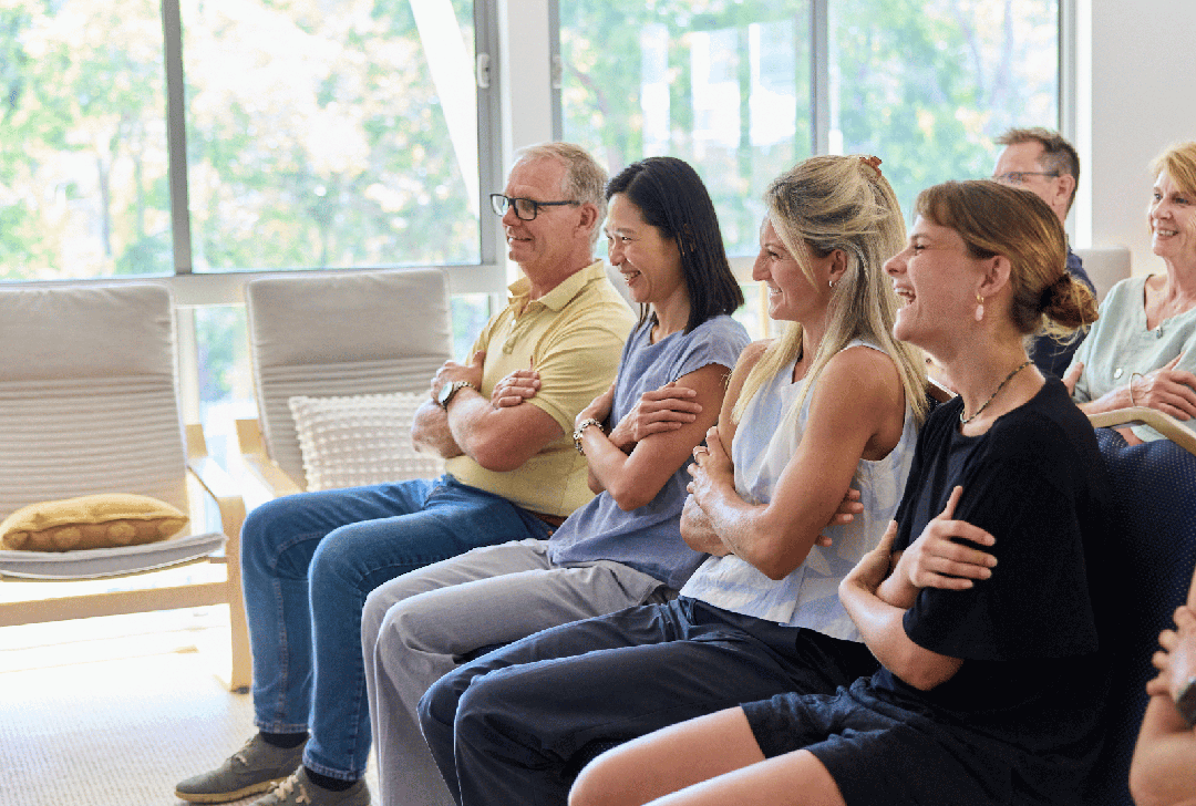People sitting on chairs indoors, laughing and smiling during a casual meeting or presentation, with large windows and greenery outside.