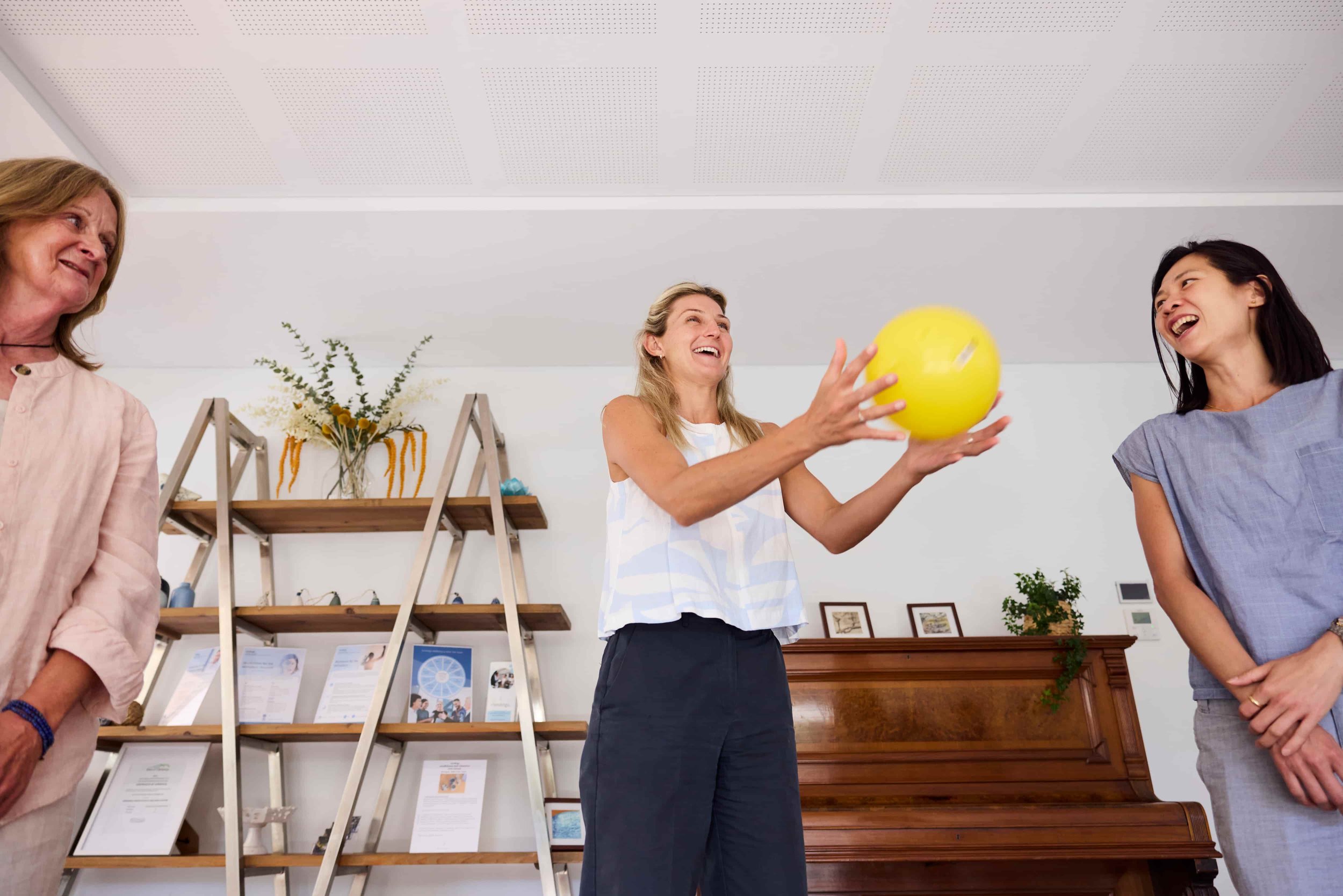 Three women enjoying a moment together, one woman in the middle is holding a yellow ball and smiling, while the other two women are smiling and laughing in a room with a piano and shelves with framed pictures and a vase with flowers in the background