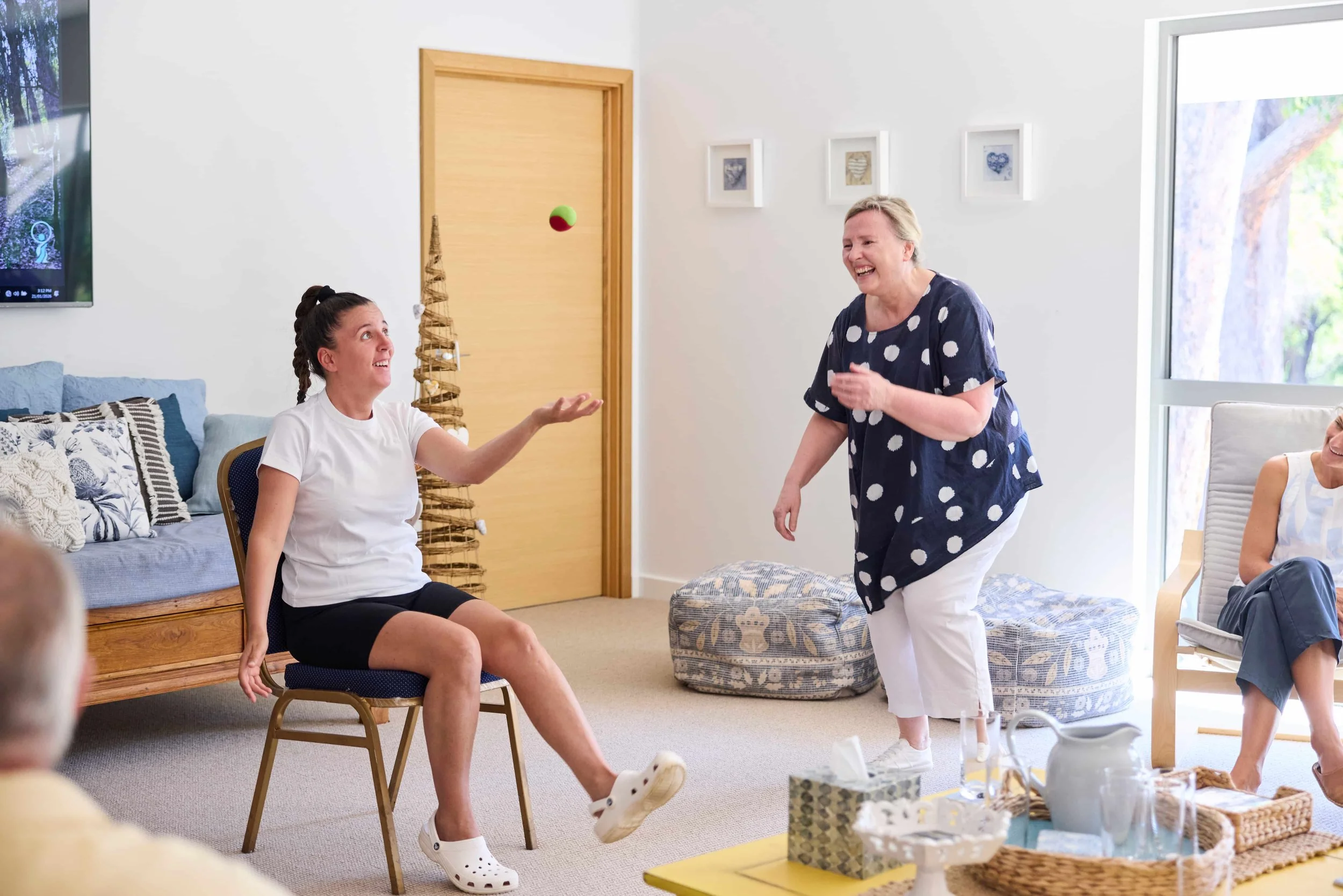 A woman in a white t-shirt and black shorts juggles a green and red ball while sitting on a chair, laughing and looking at an older woman in a navy polka dot top and white pants, who is smiling and standing in front of her. The scene is in a bright l
