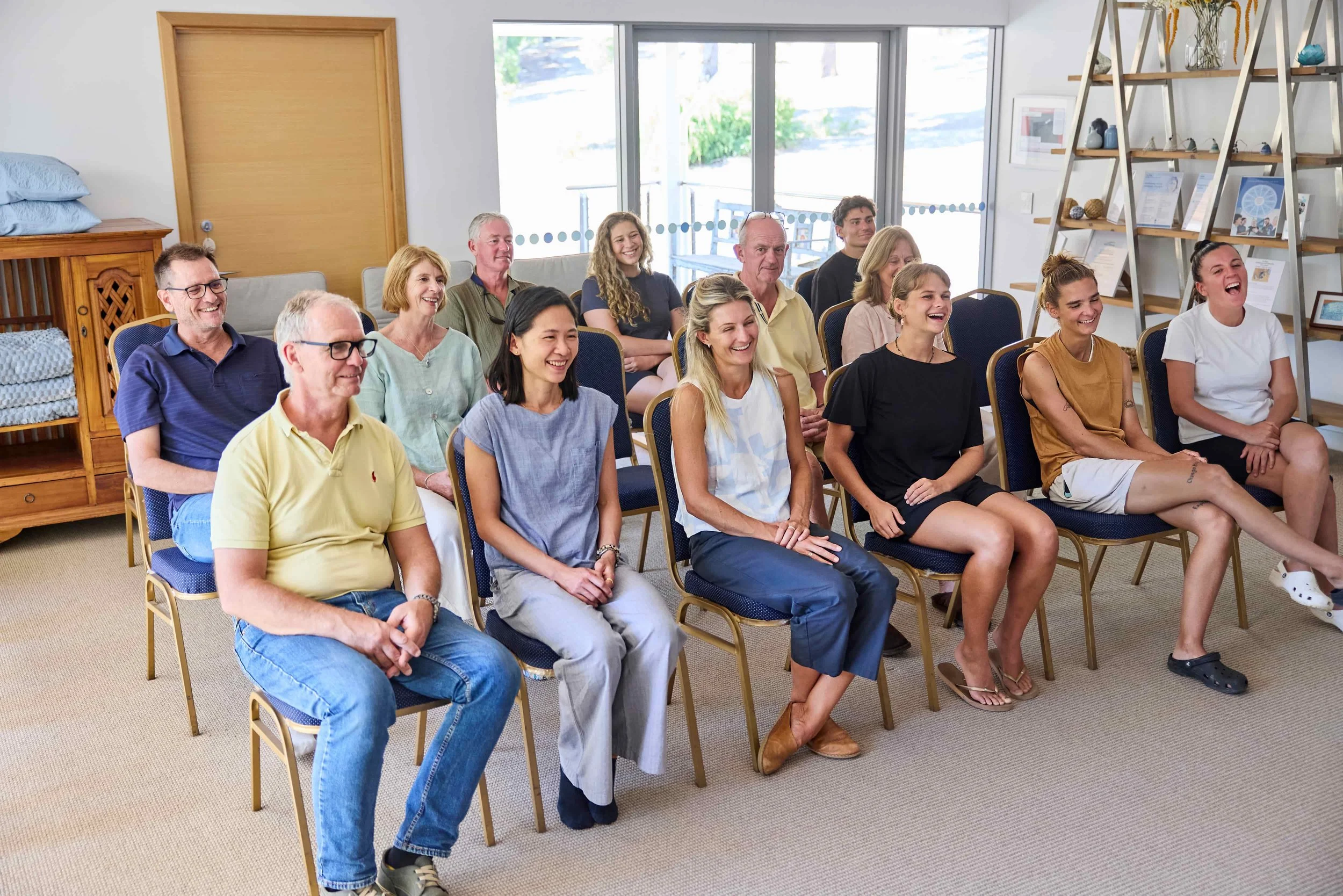 A group of people seated in a room, laughing and enjoying a presentation or event, with windows and shelves in the background.