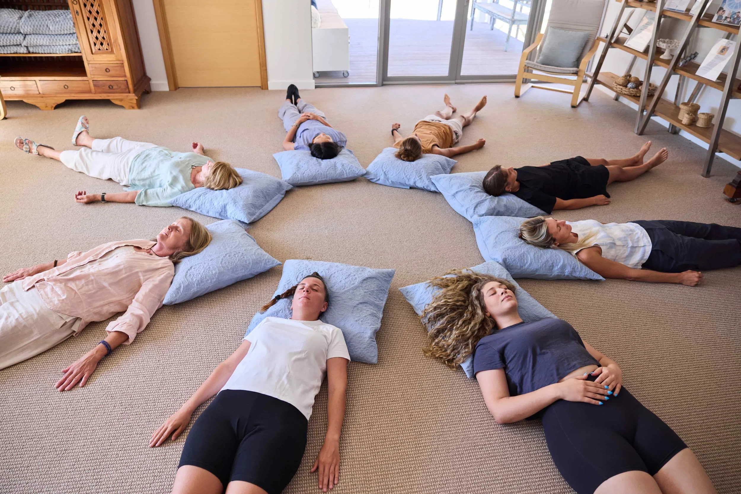 Group of nine people lying on the carpeted floor, resting on pillows in a semi-circle with eyes closed in a peaceful state.