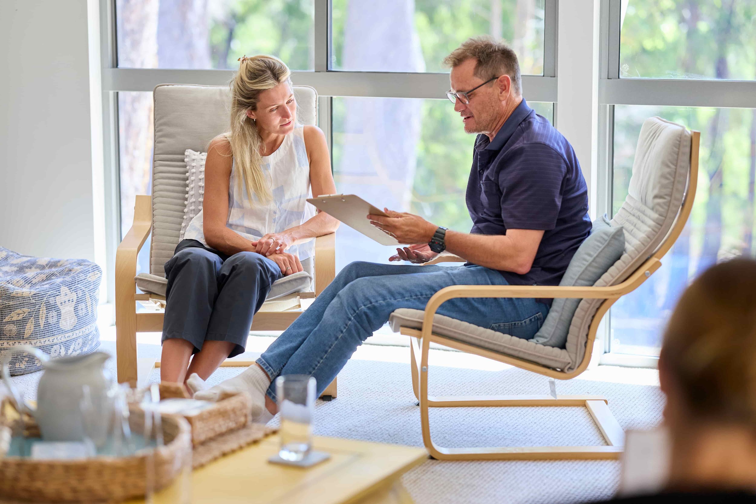 A woman and a man having a conversation in a modern, brightly lit room with large windows showing trees outside. The woman is sitting on a cushioned armchair and the man is on a wooden chair with a cushion, holding a clipboard.