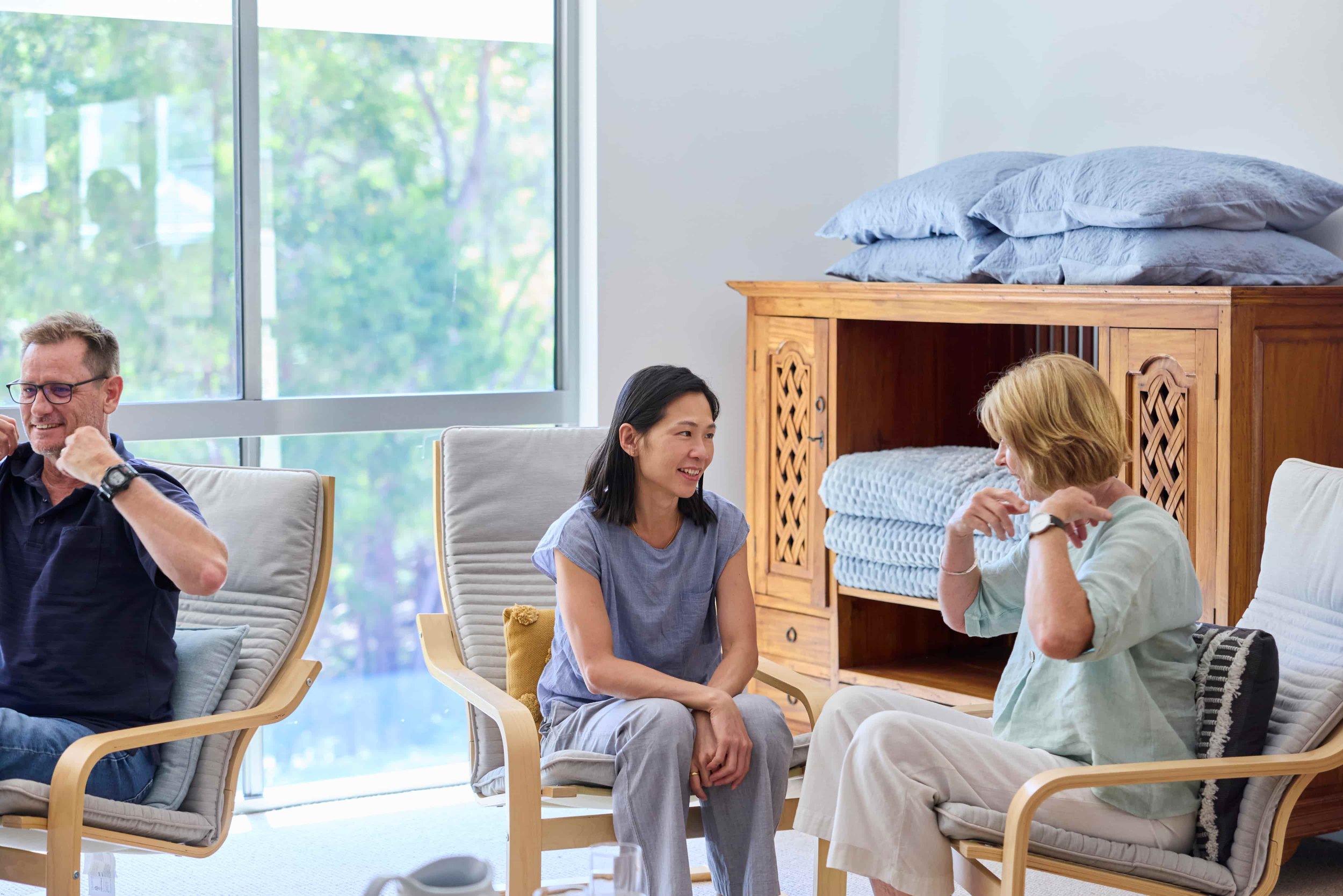 Three people sitting in a bright, modern room near a large window, engaged in conversation; a man on the left, a woman in the middle, and another woman on the right.