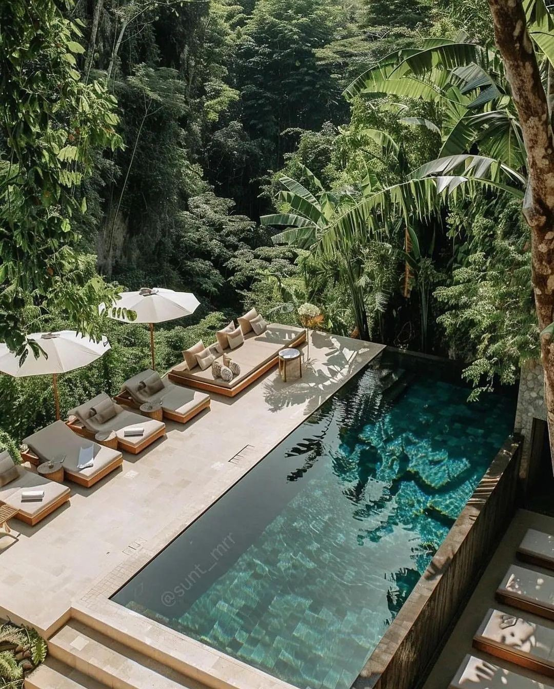 Luxury poolside area with lounge chairs, umbrellas, and a seating area surrounded by lush tropical foliage.