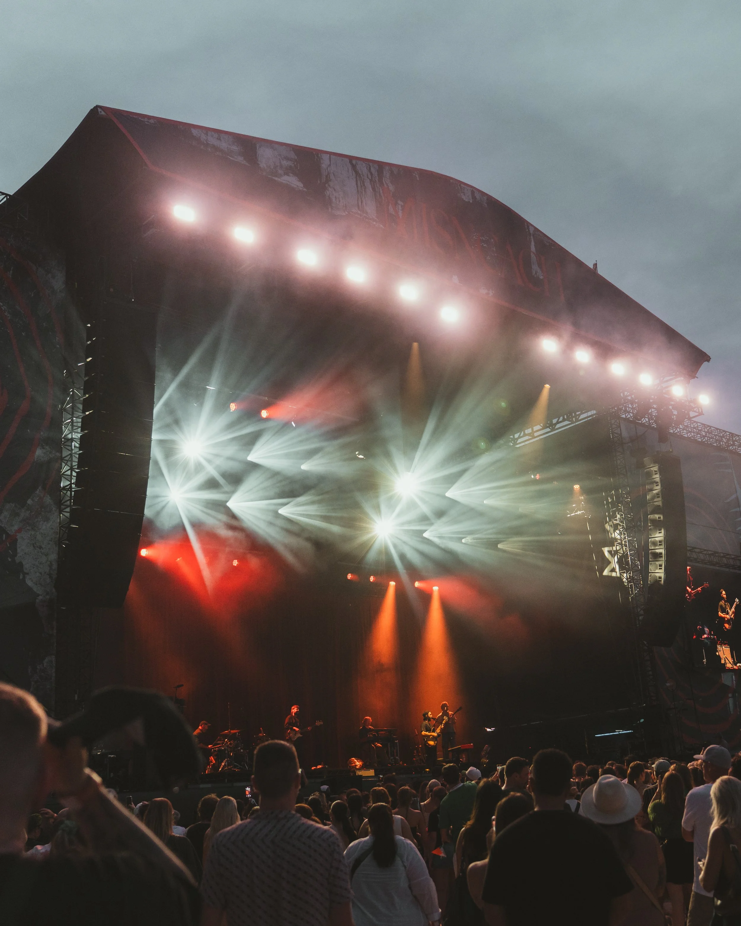 Outdoor concert stage with bright lights and performing band, audience standing and watching during evening.