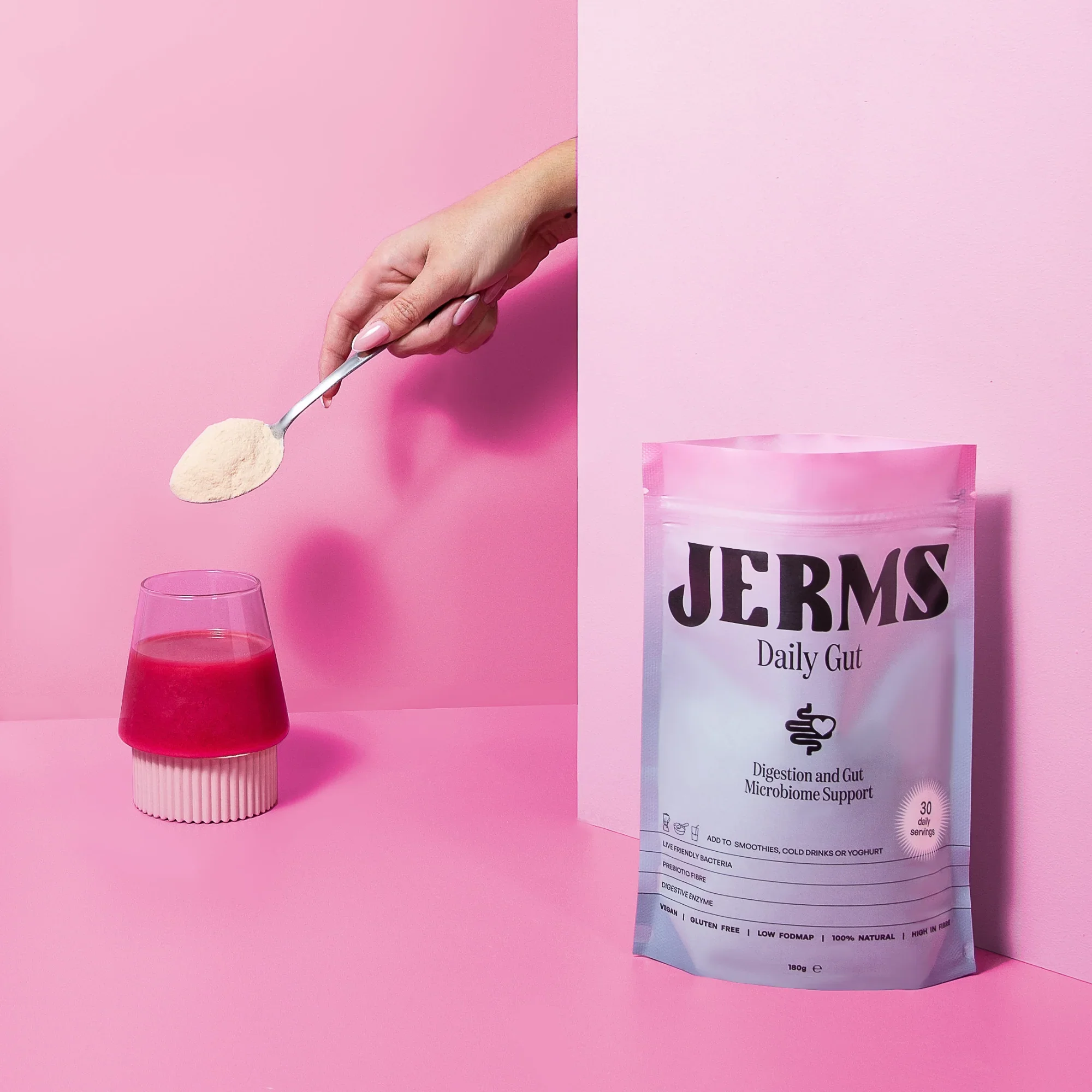Hand holding a spoon of powder over a glass of pink liquid, with a pink background and a bag labeled 'JERMS Daily Gut' probiotic supplement beside it.