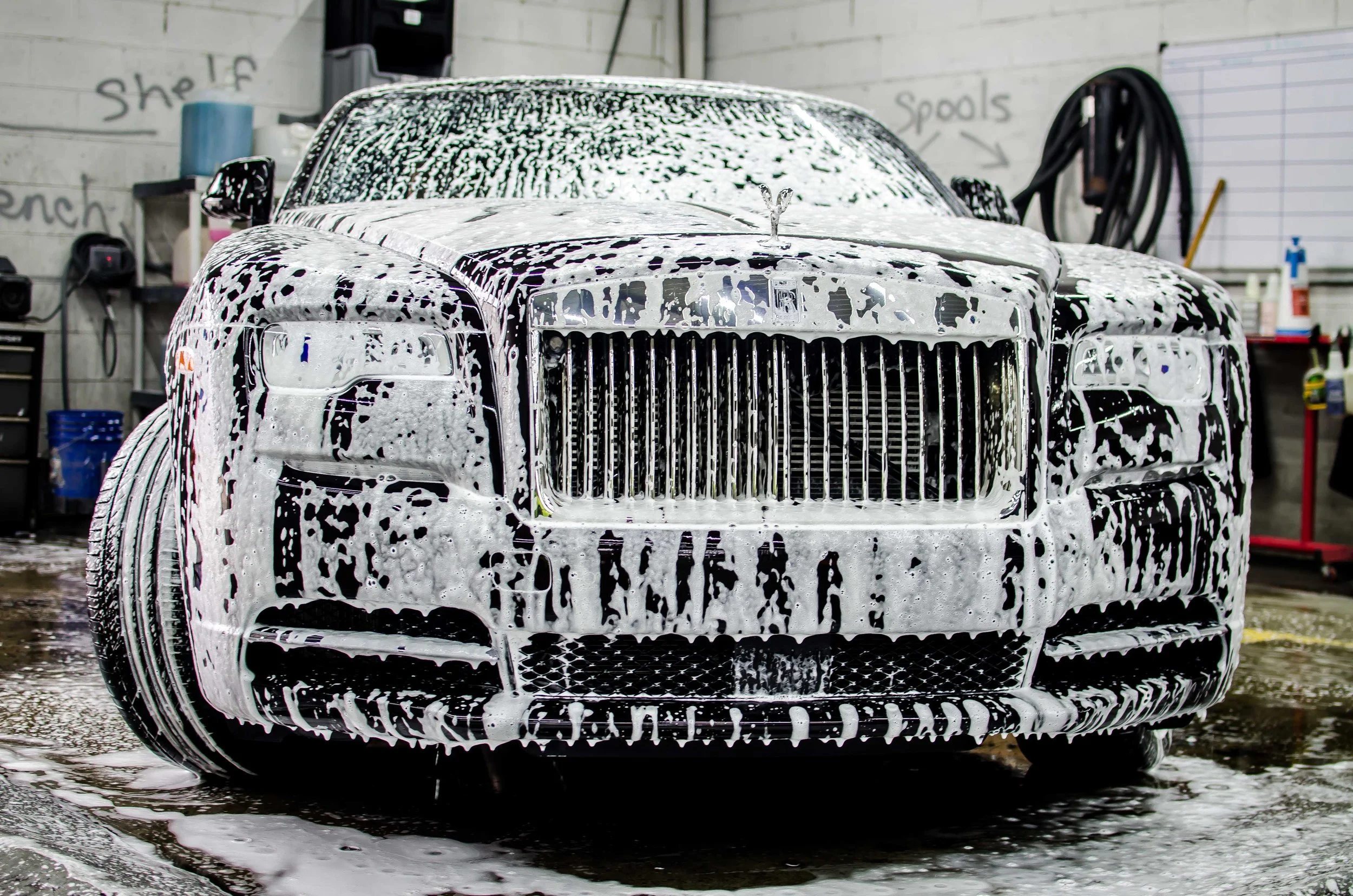 Front view of a black luxury car covered in white soap suds during a washing process inside a garage or car wash station.