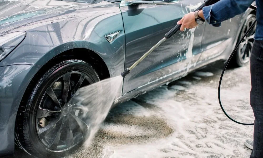 Person washing a gray electric car with a pressure washer, soap and water spraying on the side of the vehicle.