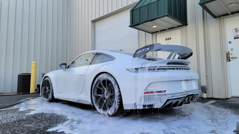 White sports car with a large rear spoiler being washed outside a building, with soap and water running off.