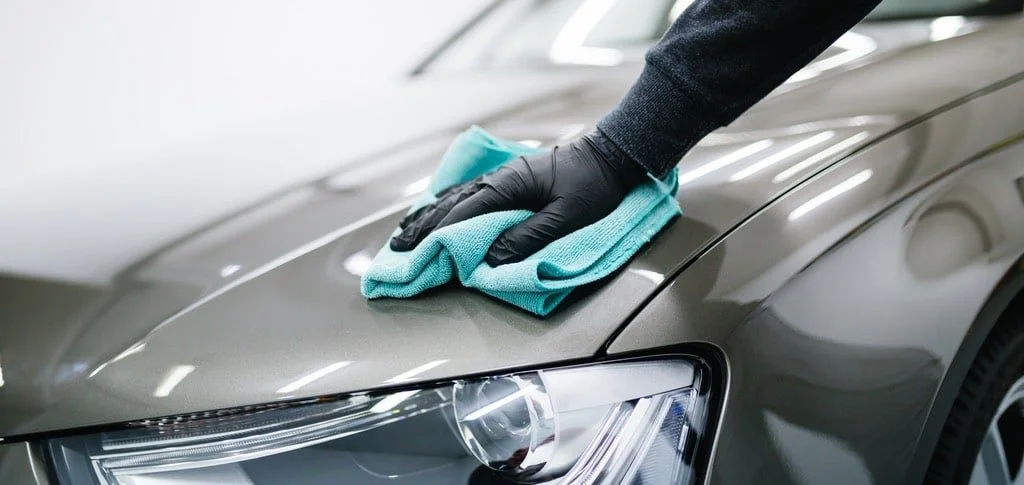 A person wearing black gloves cleaning a gray car with a blue cloth.