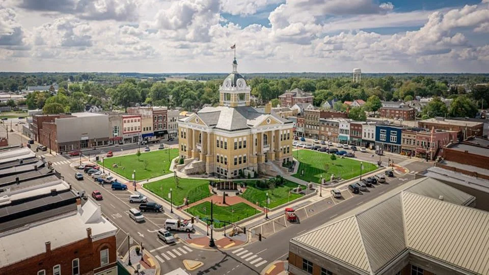 A historic courthouse with a dome and clock tower situated in the center of a small town, surrounded by streets, cars, and colorful shops.