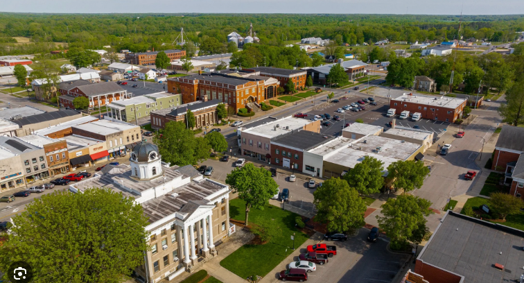 Aerial view of a small town with historic buildings, parking lots, trees, and surrounding green fields.