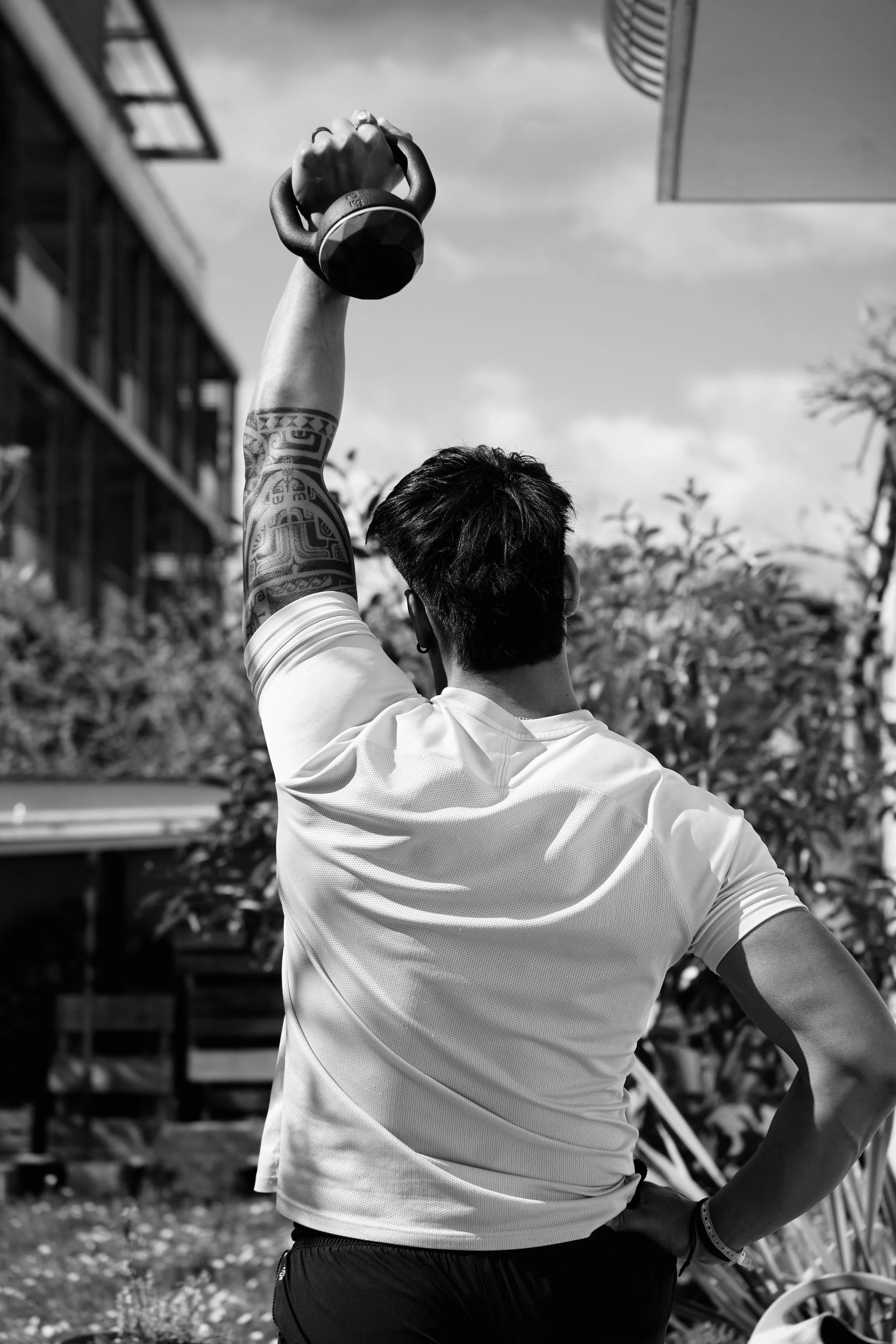 Une personne vue de dos, tenant un kettlebell levé en l'air, portant un t-shirt blanc et un bracelet, avec un tatouage sur le bras, en plein air avec des bâtiments et des arbres en arrière-plan.