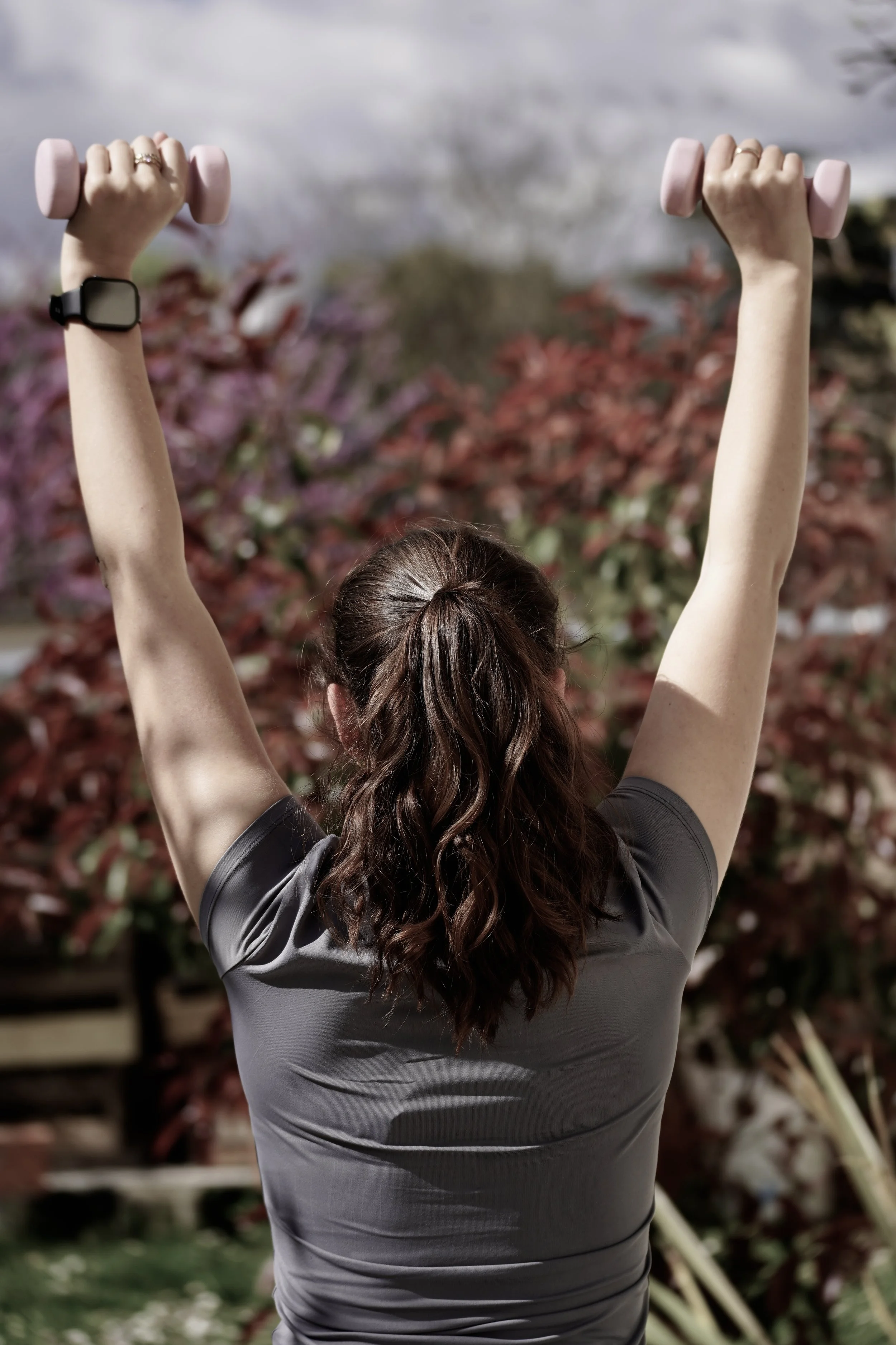 Femme faisant de l'exercice en levant des haltères roses dans un jardin, vue de dos.