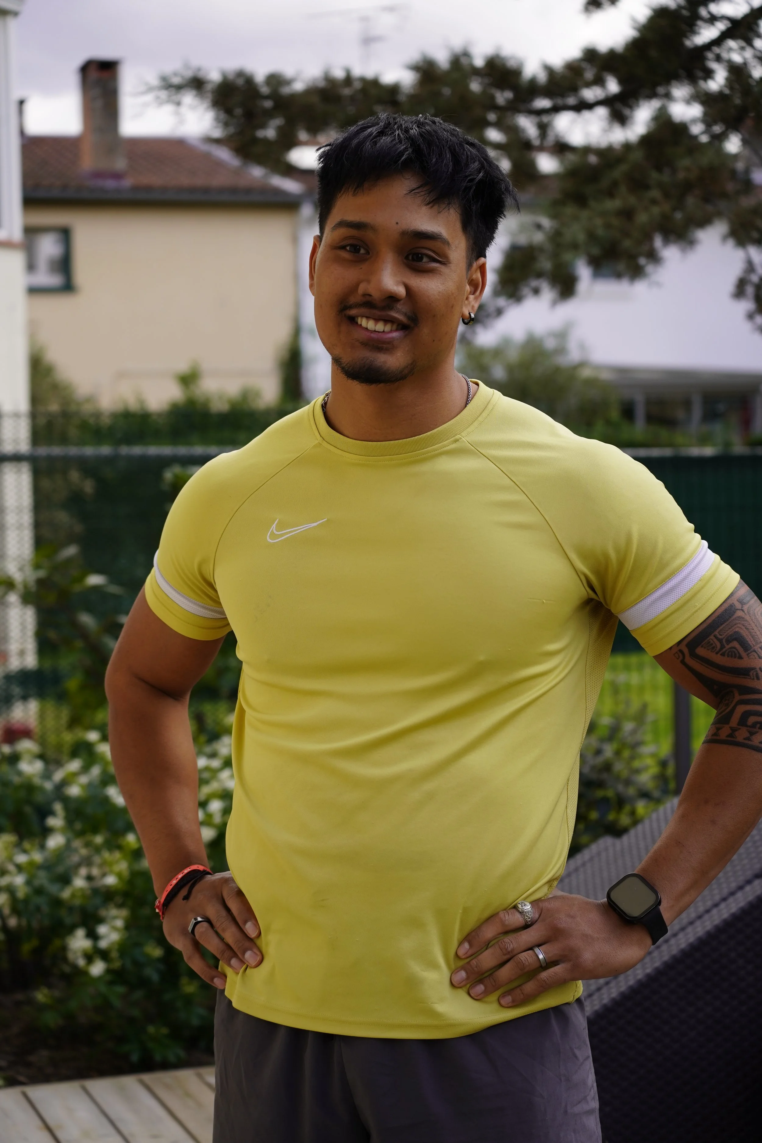 Un homme souriant portant un t-shirt jaune et un smartwatch, posant avec les mains sur les hanches dans un jardin avec des plantes et une maison en arrière-plan.