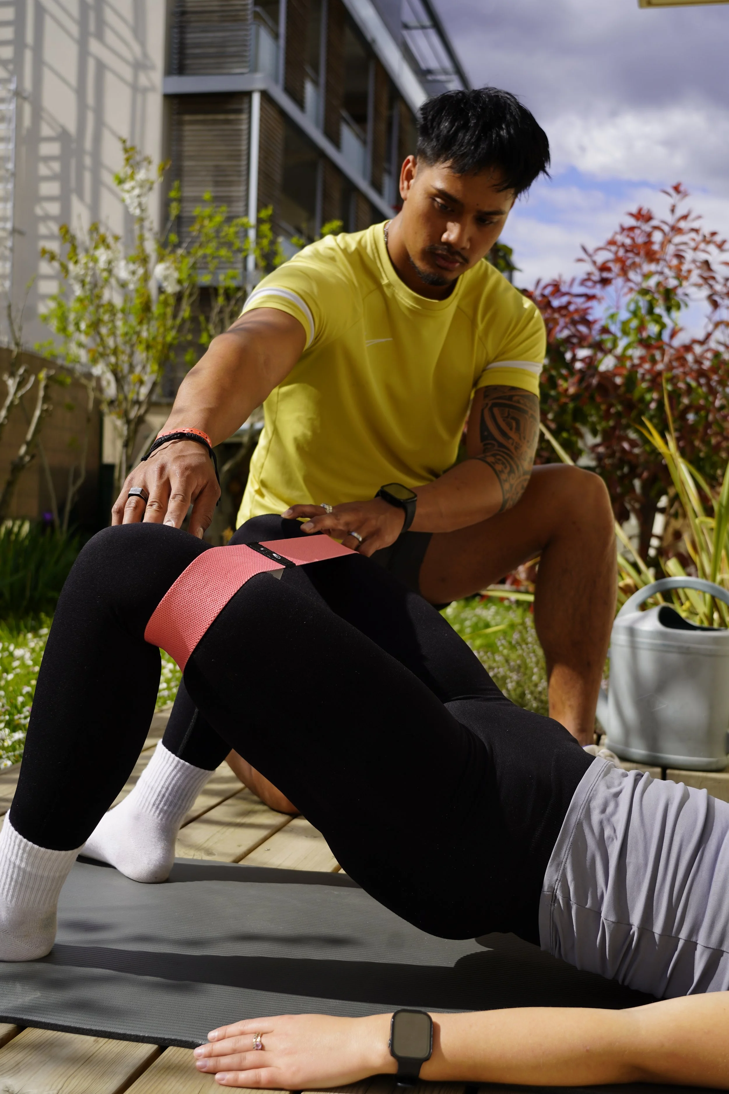 Un homme aide une autre personne à faire de la physiothérapie ou des exercices de rééducation sur un tapis de yoga dans un jardin. L'homme porte un t-shirt jaune et une montre connectée.