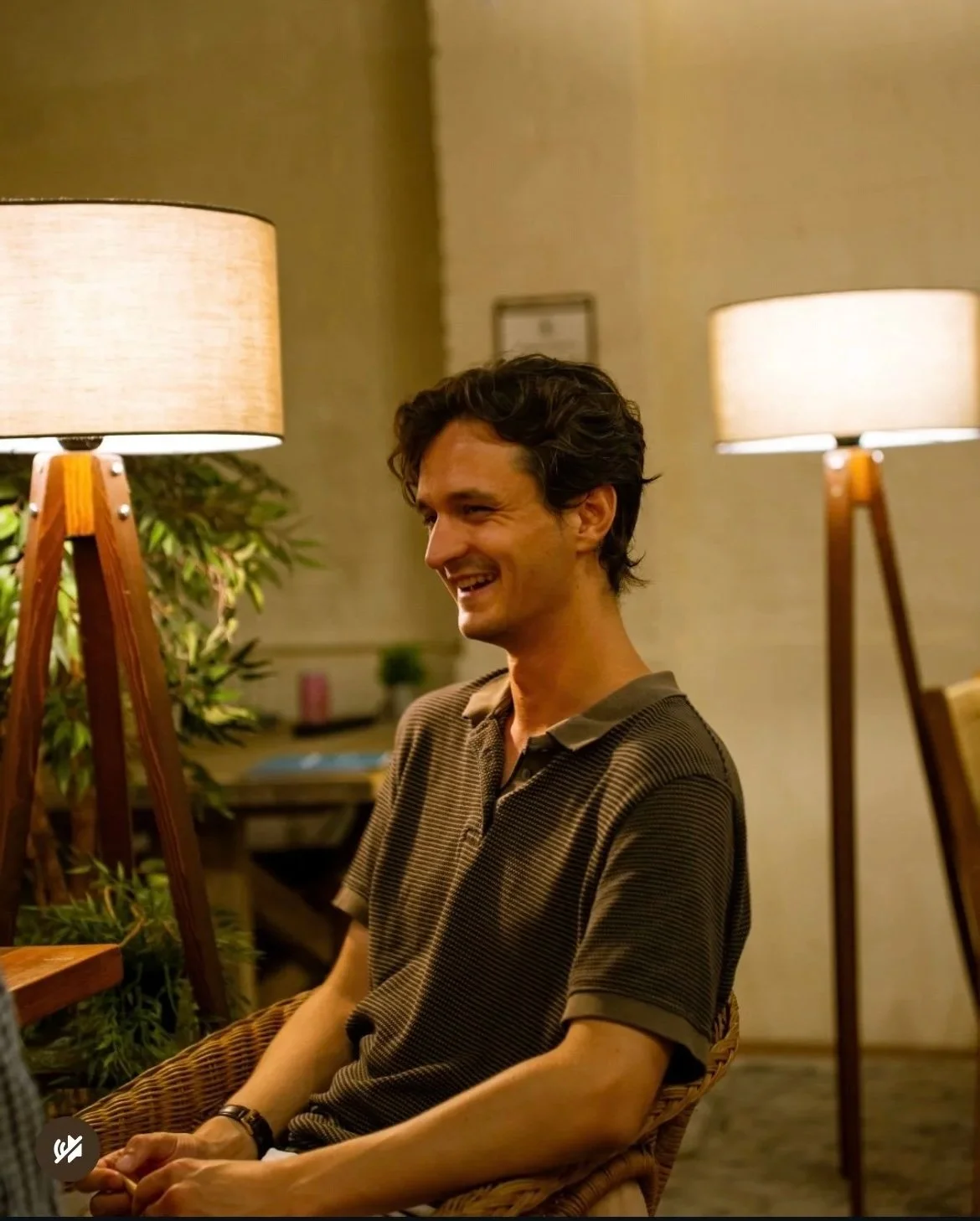 A man with dark, wavy hair smiling while sitting in a wicker chair in a warmly lit room with two standing lamps and green plants.